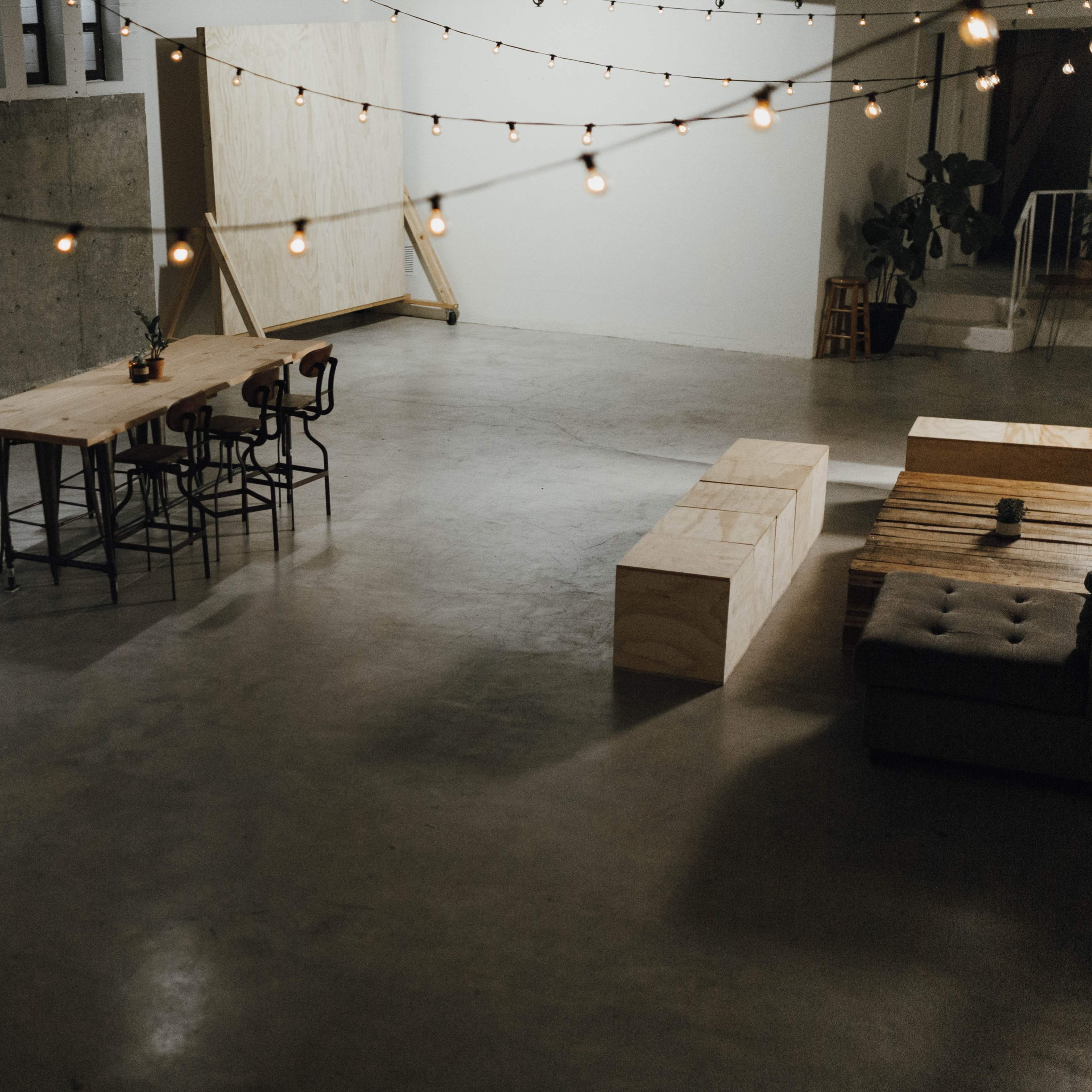 A spacious, minimalist room with a long wooden table, chairs, and string lights hanging overhead.