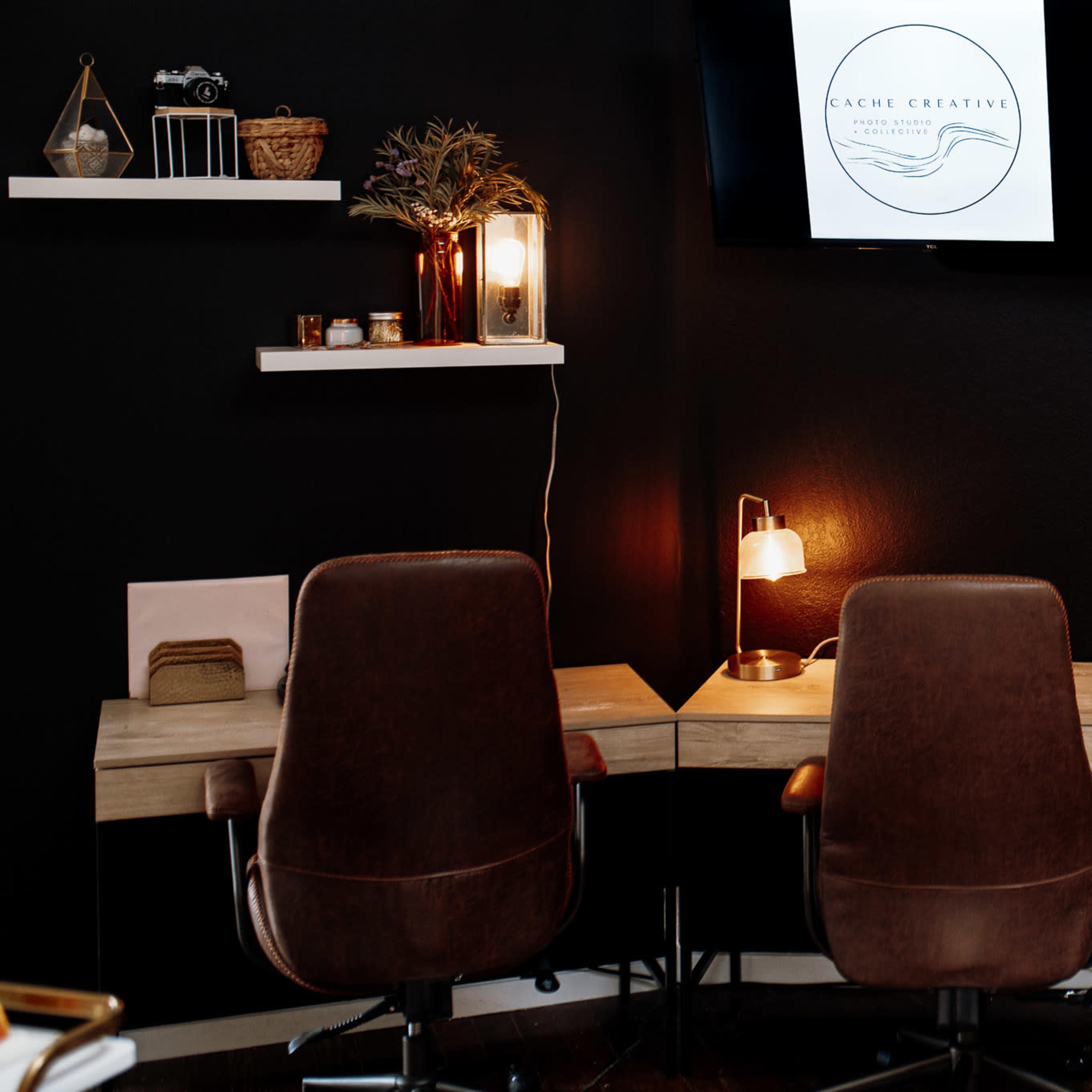 A modern workspace featuring two brown office chairs facing a small wooden desk against a dark wall, with shelves displaying decorative items and a logo on a screen.