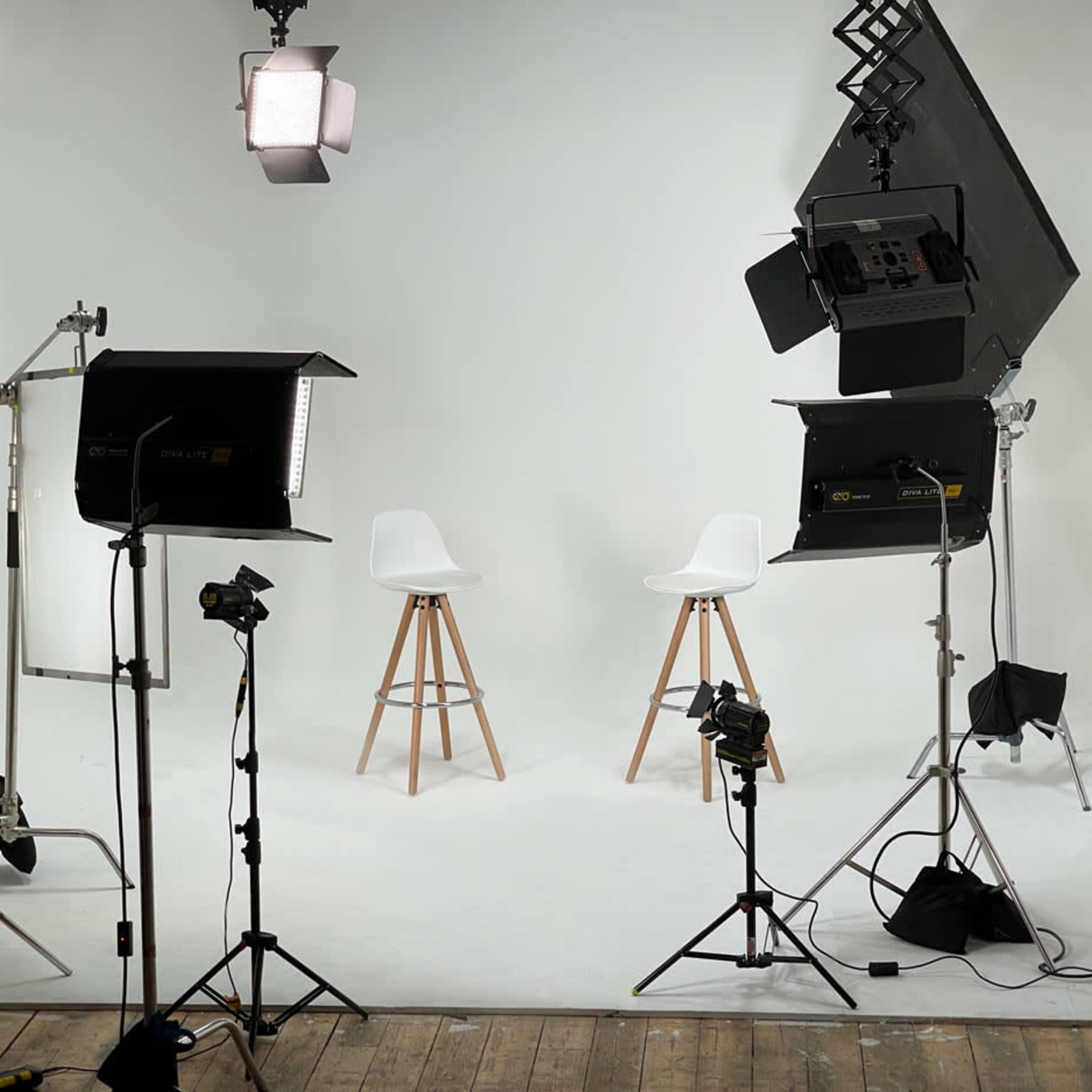 A photography studio setup features two white chairs on wooden stools, surrounded by various lighting equipment against a plain backdrop.
