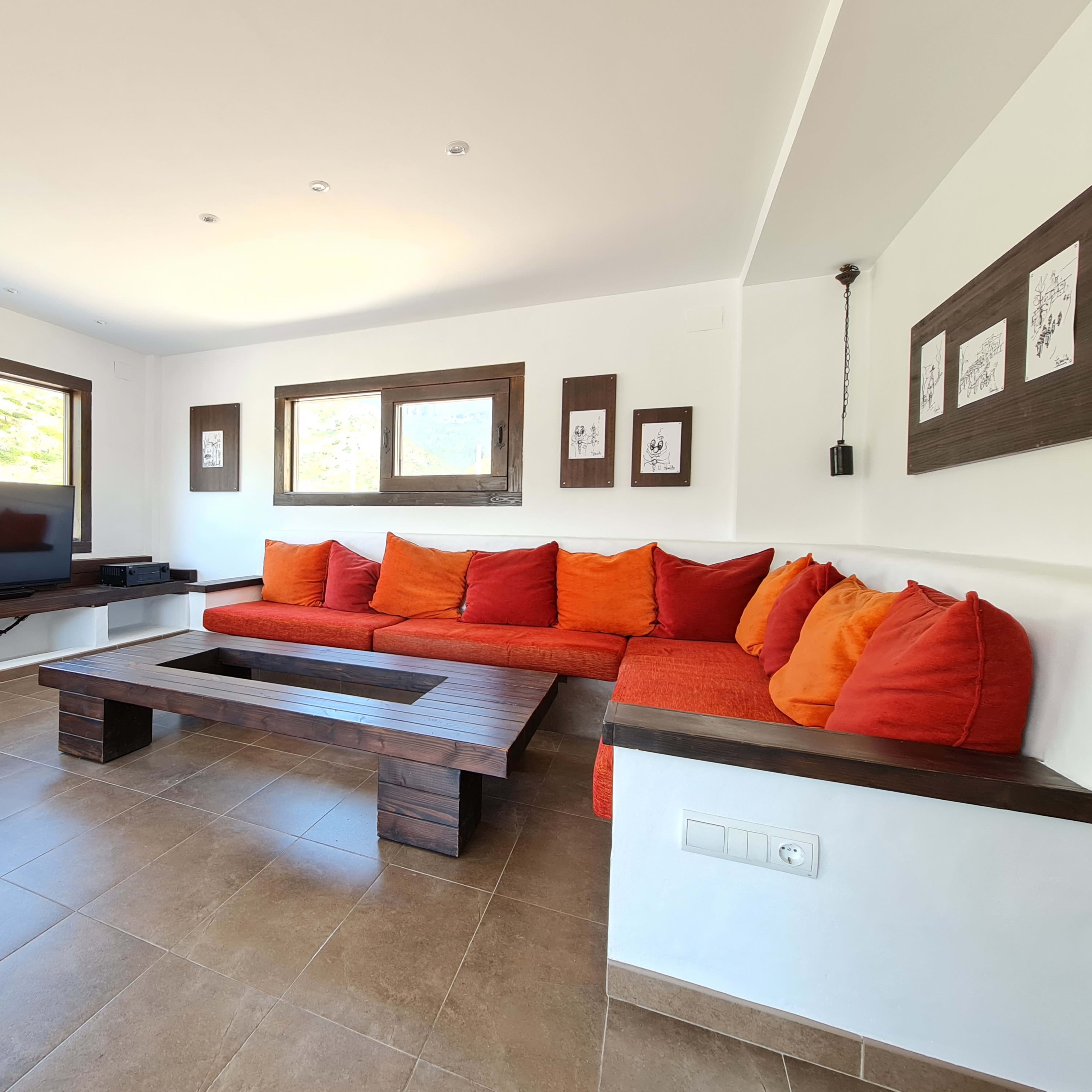 A modern living room with a large orange sectional sofa, a wooden coffee table, and a TV mounted on the wall, surrounded by large windows letting in natural light.