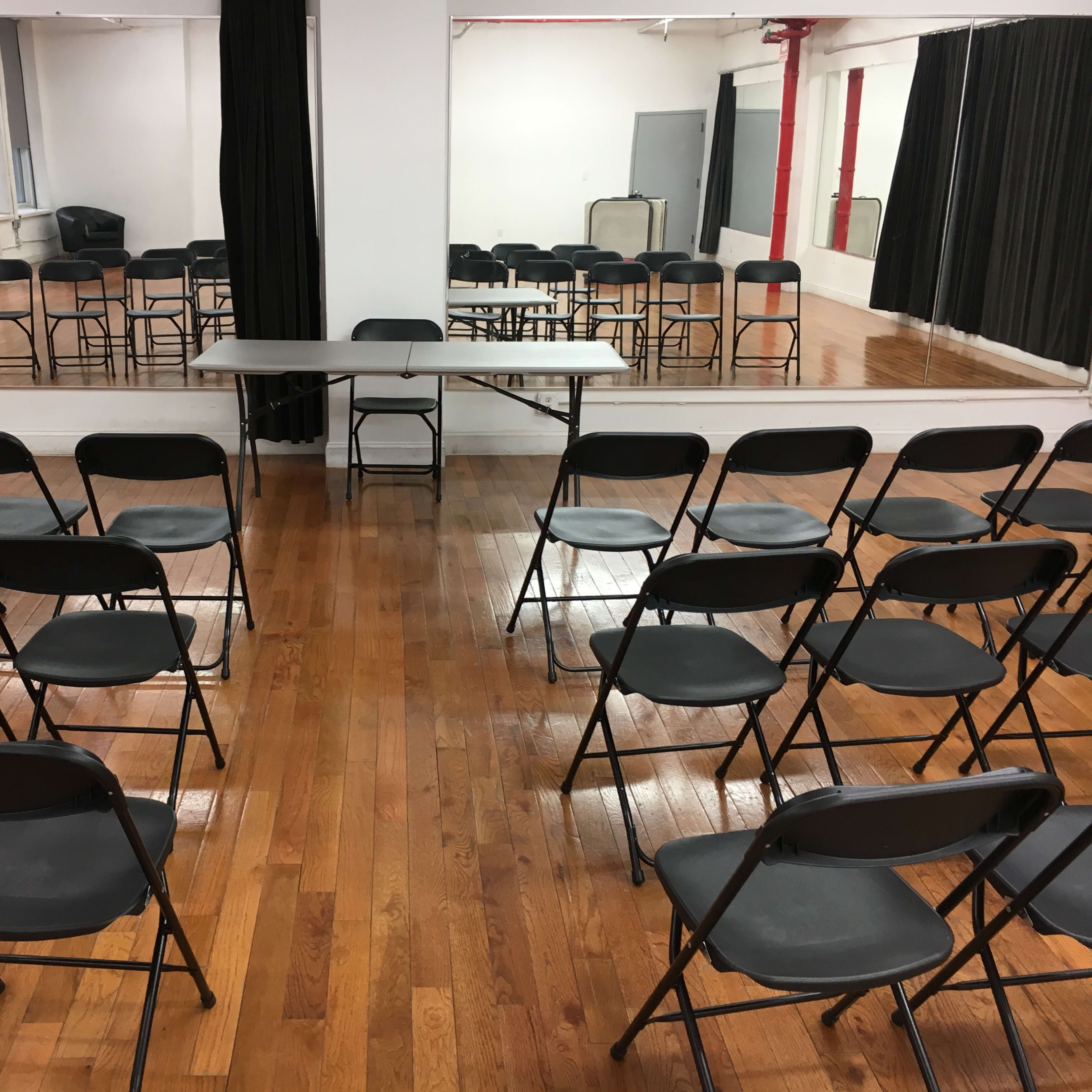 A room with wooden flooring, where black folding chairs are arranged in rows facing a table, with mirrors and additional chairs visible in the background.