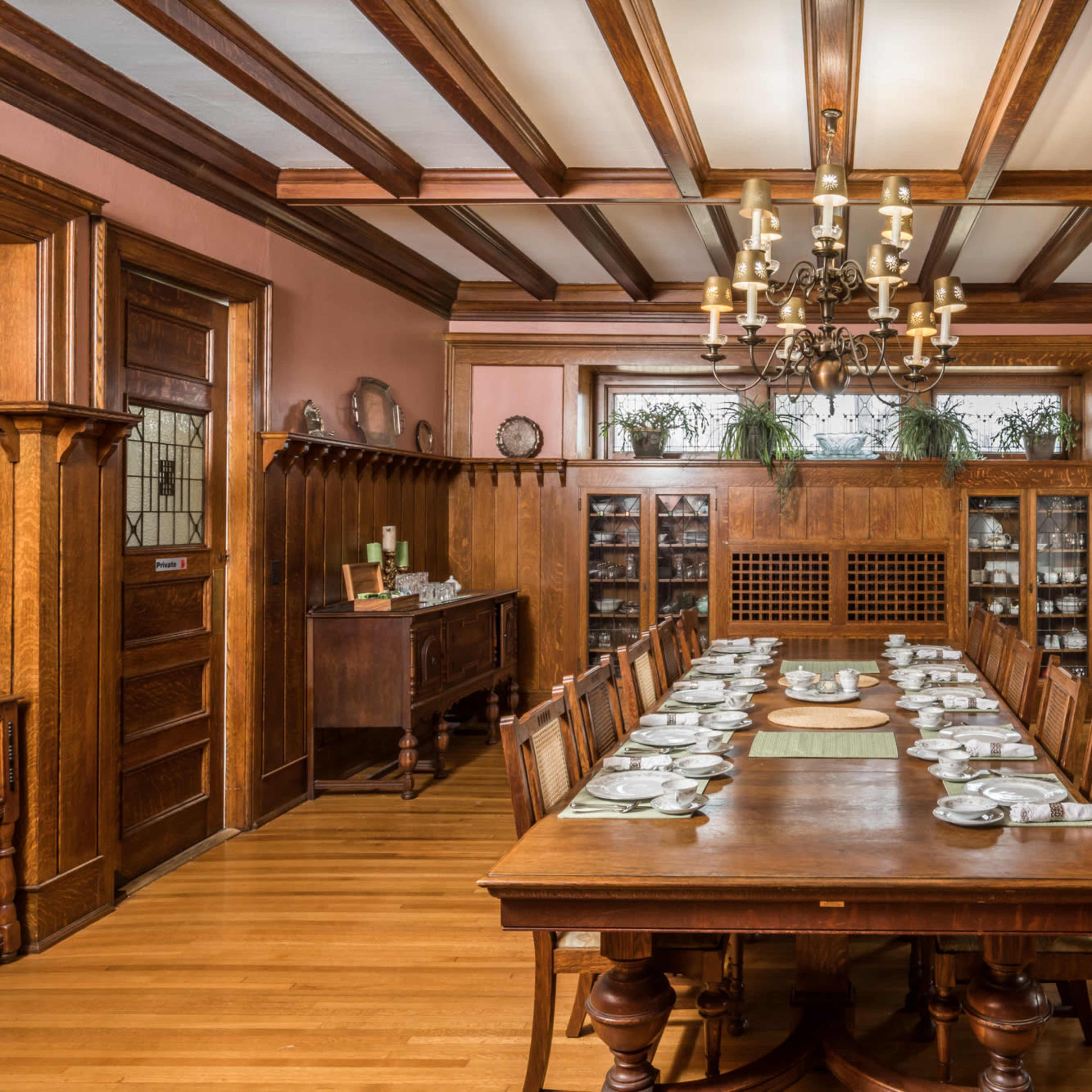 A formal dining room with a long wooden table set for a meal, surrounded by wooden chairs, and featuring chandeliers and decorative woodwork.