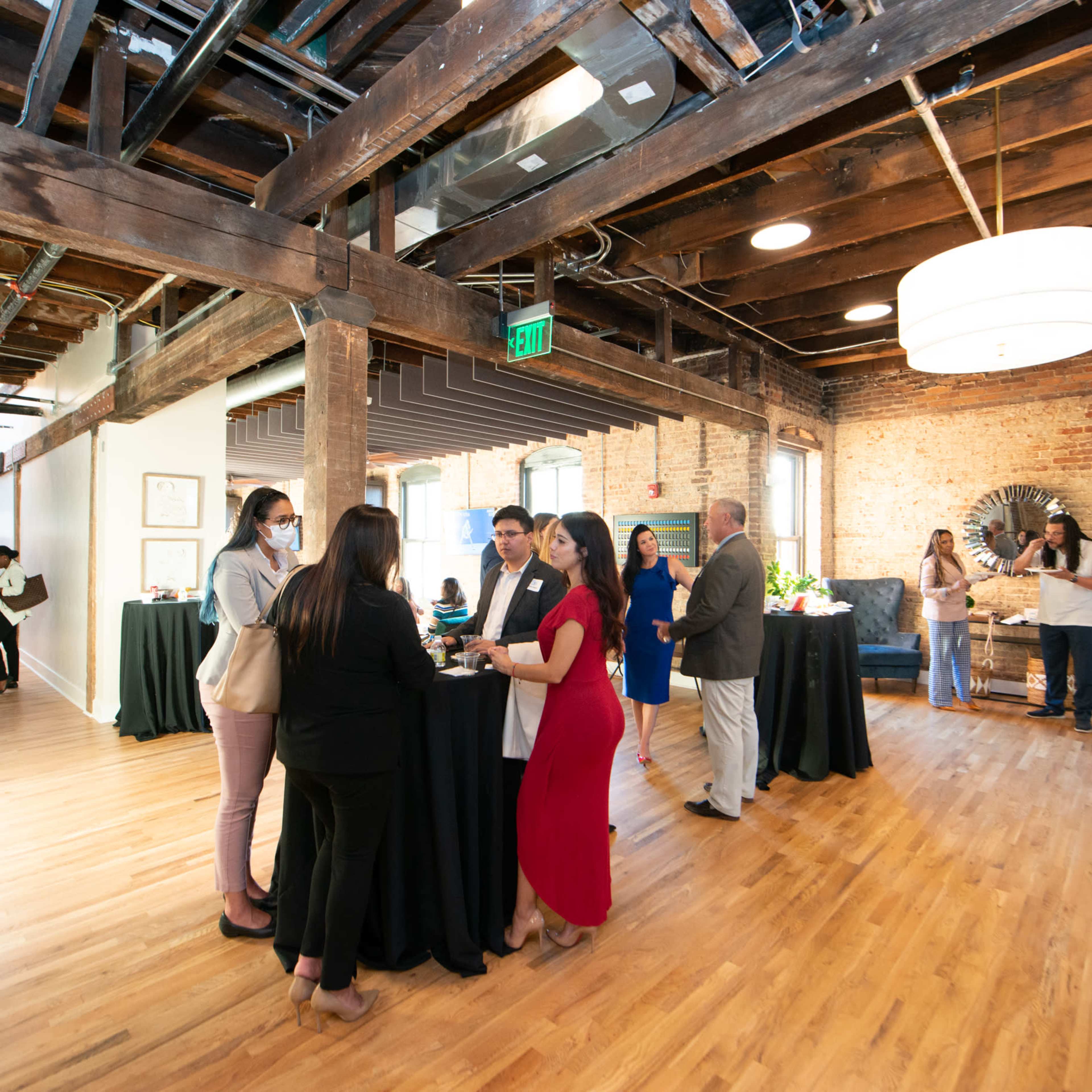 A group of people engages in conversation at a gathering in a well-lit, rustic space with exposed wooden beams and a mix of modern and vintage decor.