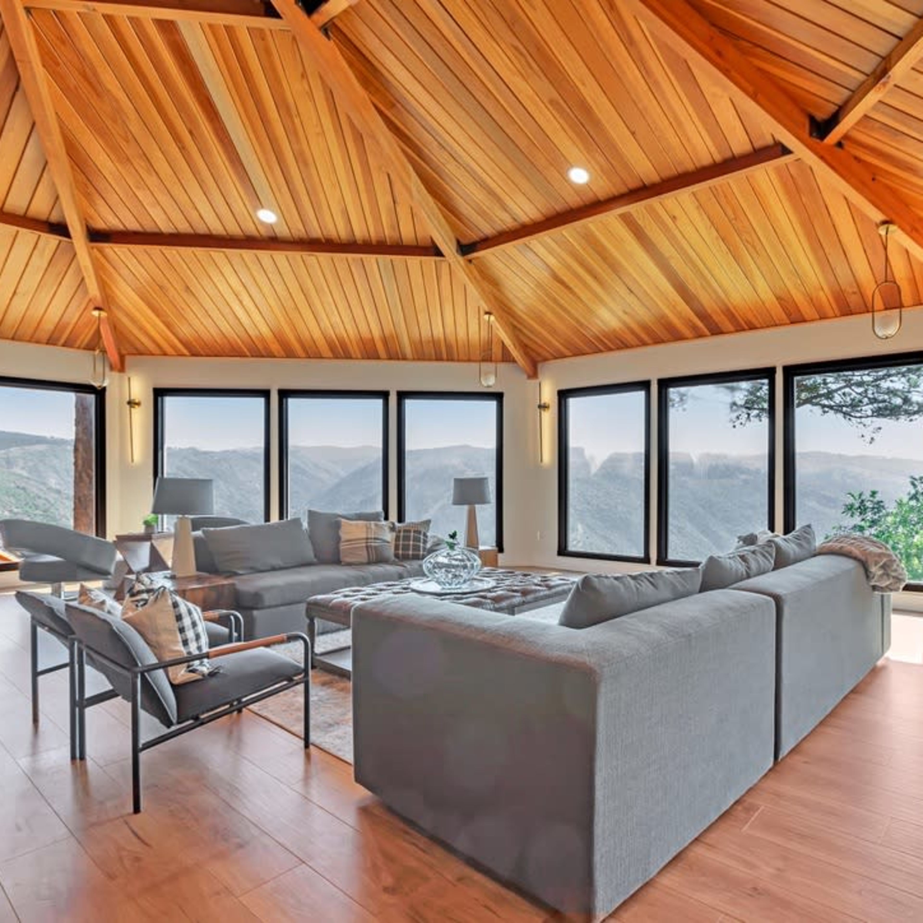 A spacious living room with a high wooden ceiling and large windows overlooking a mountainous landscape.