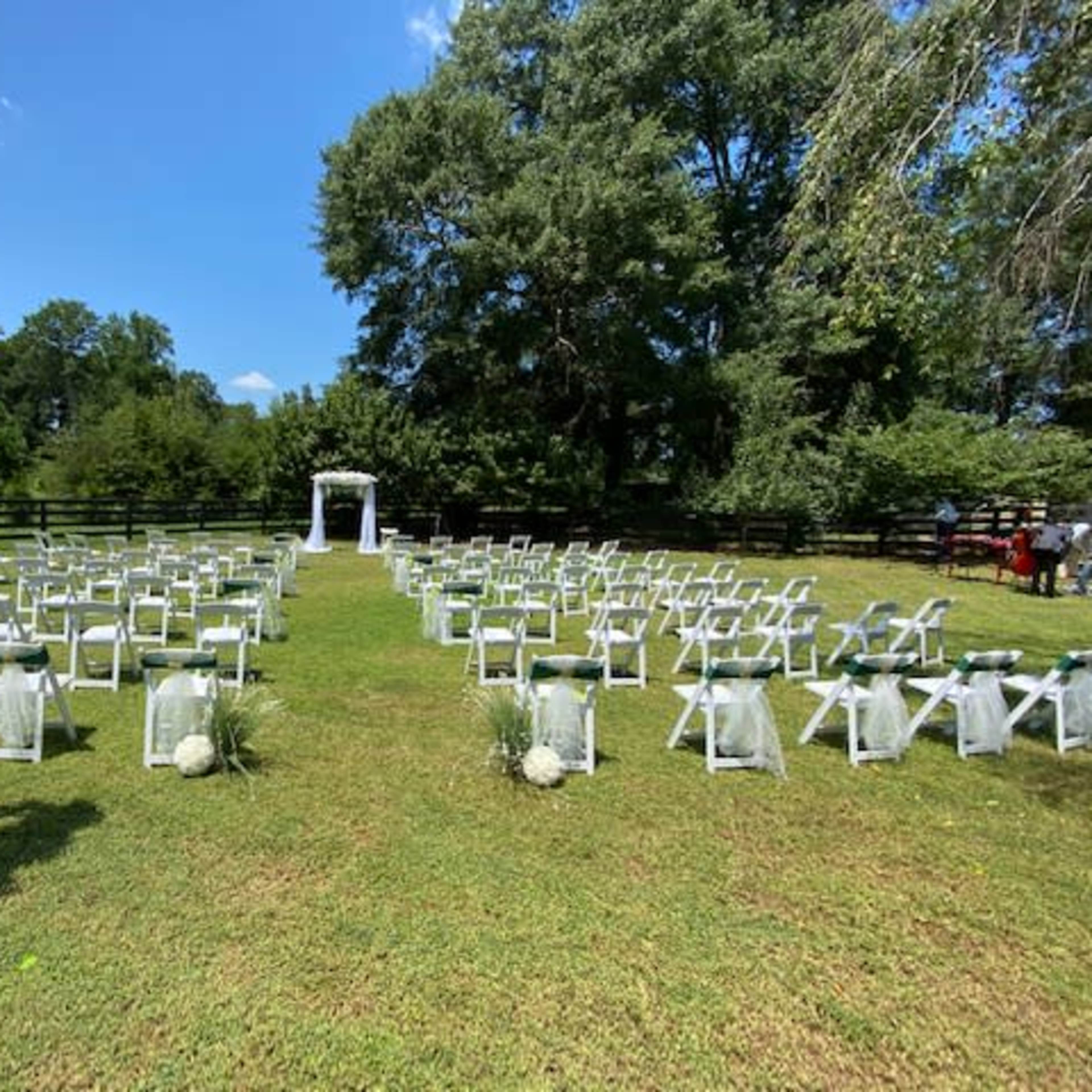 A wedding ceremony setup features white chairs arranged in rows on a grassy field, facing an arch adorned with flowers and greenery.
