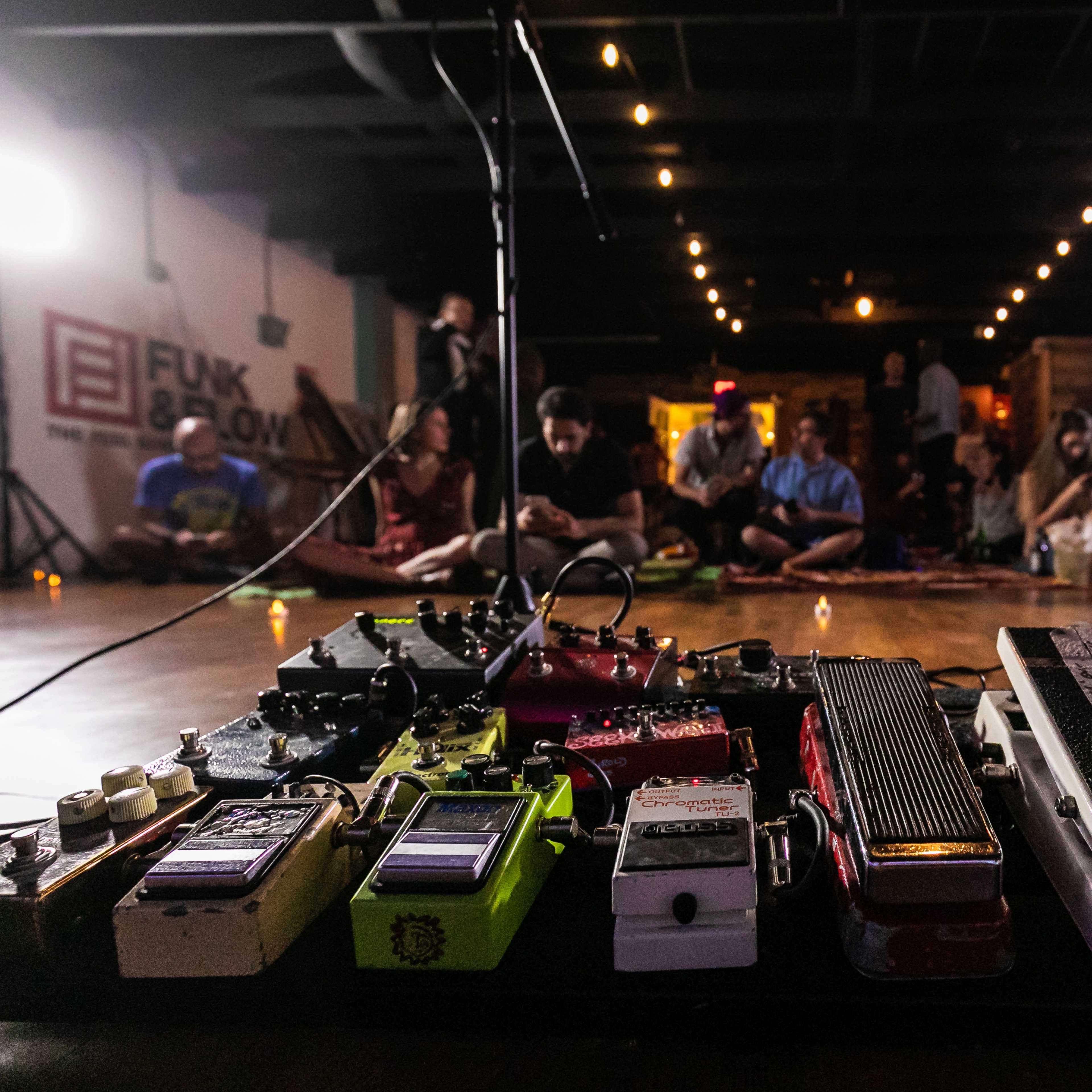 A collection of guitar effects pedals is prominently displayed in the foreground, with an audience seated in a dimly lit venue in the background.