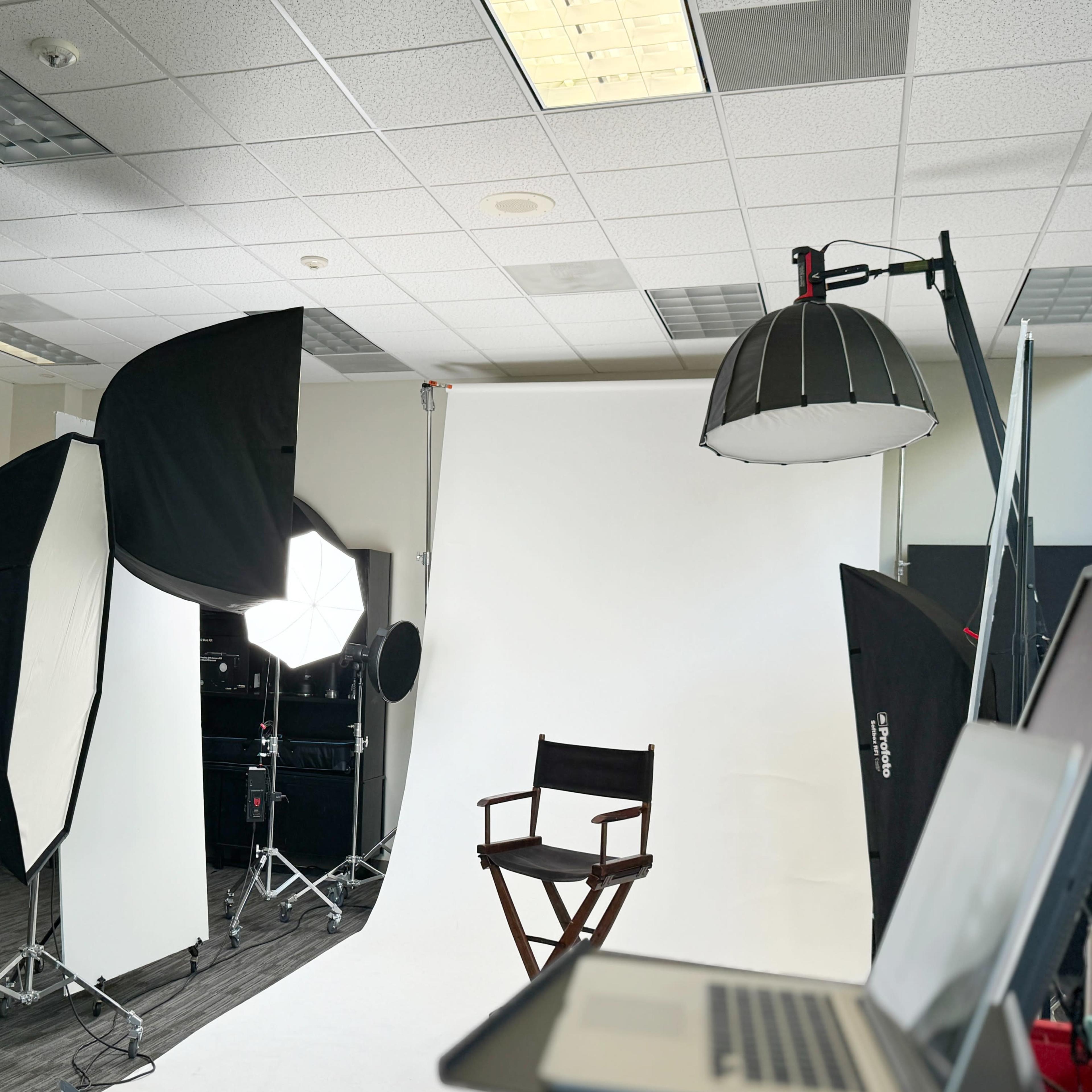 A photography studio is set up with various lights and reflectors aimed at a blank backdrop, featuring a director's chair in the center.