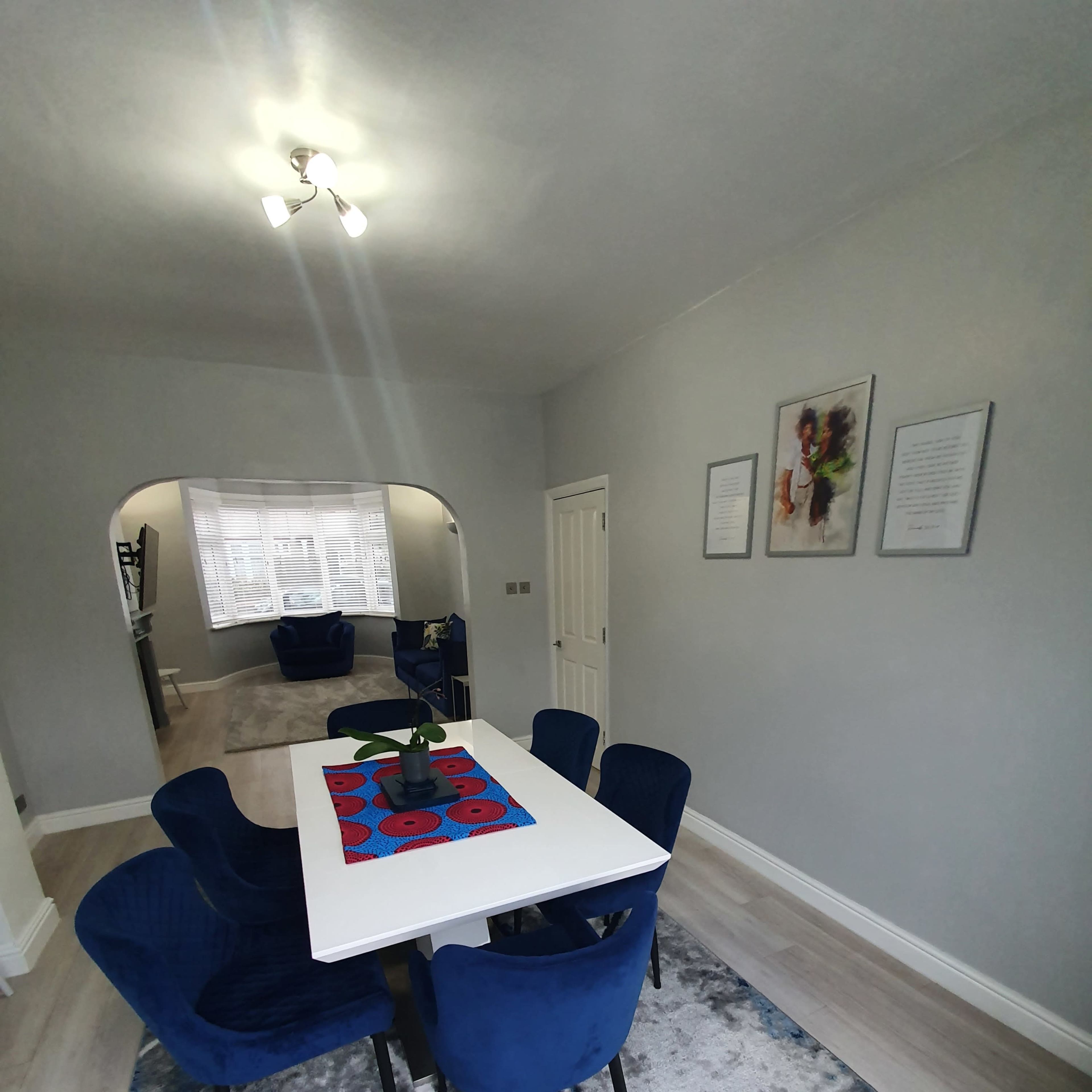 A dining area features a white table with a red and black centerpiece surrounded by blue upholstered chairs, with light gray walls and a light-colored floor.