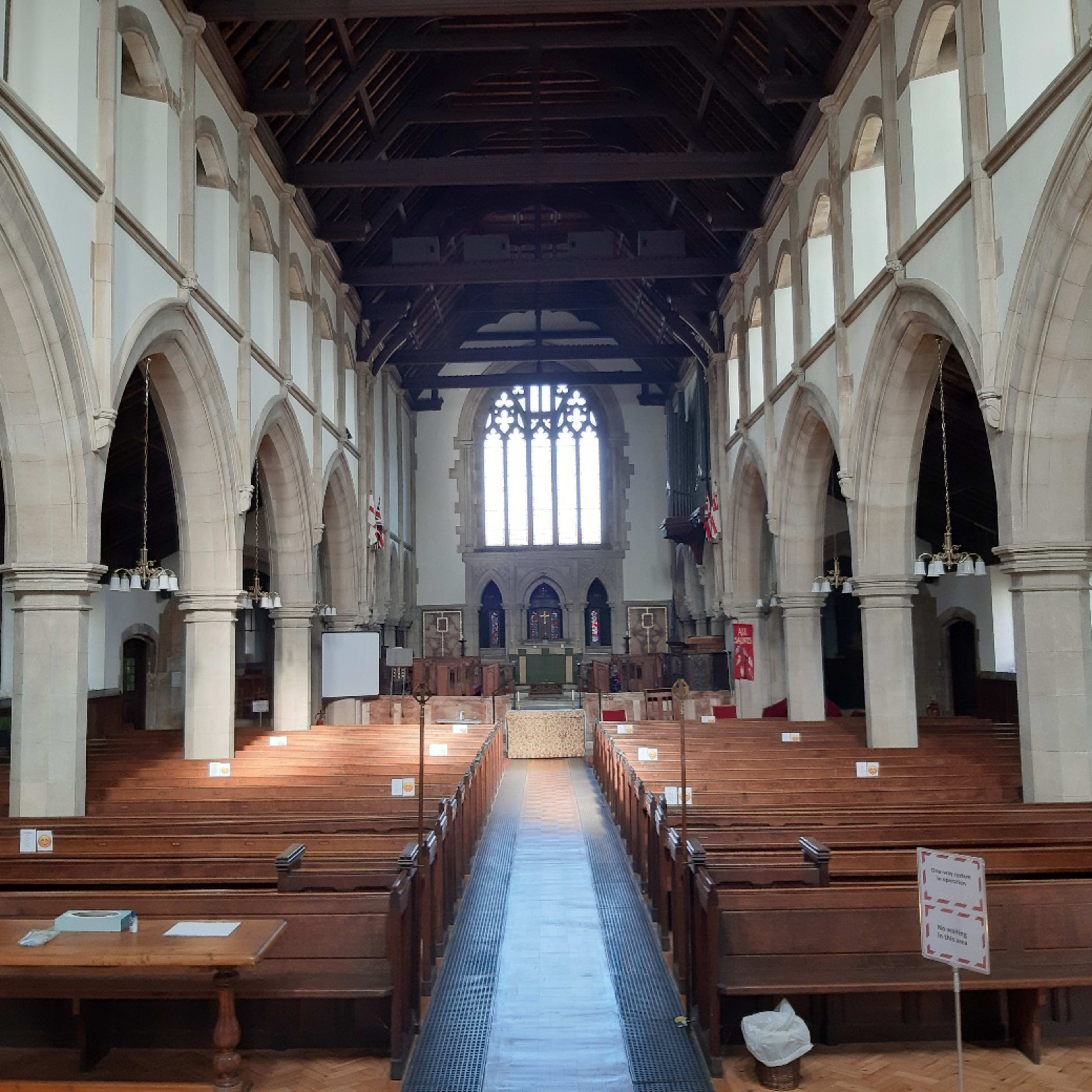 The interior of a large church featuring wooden pews arranged in rows, arched stone columns, and a high, beamed ceiling with a large stained glass window at one end.