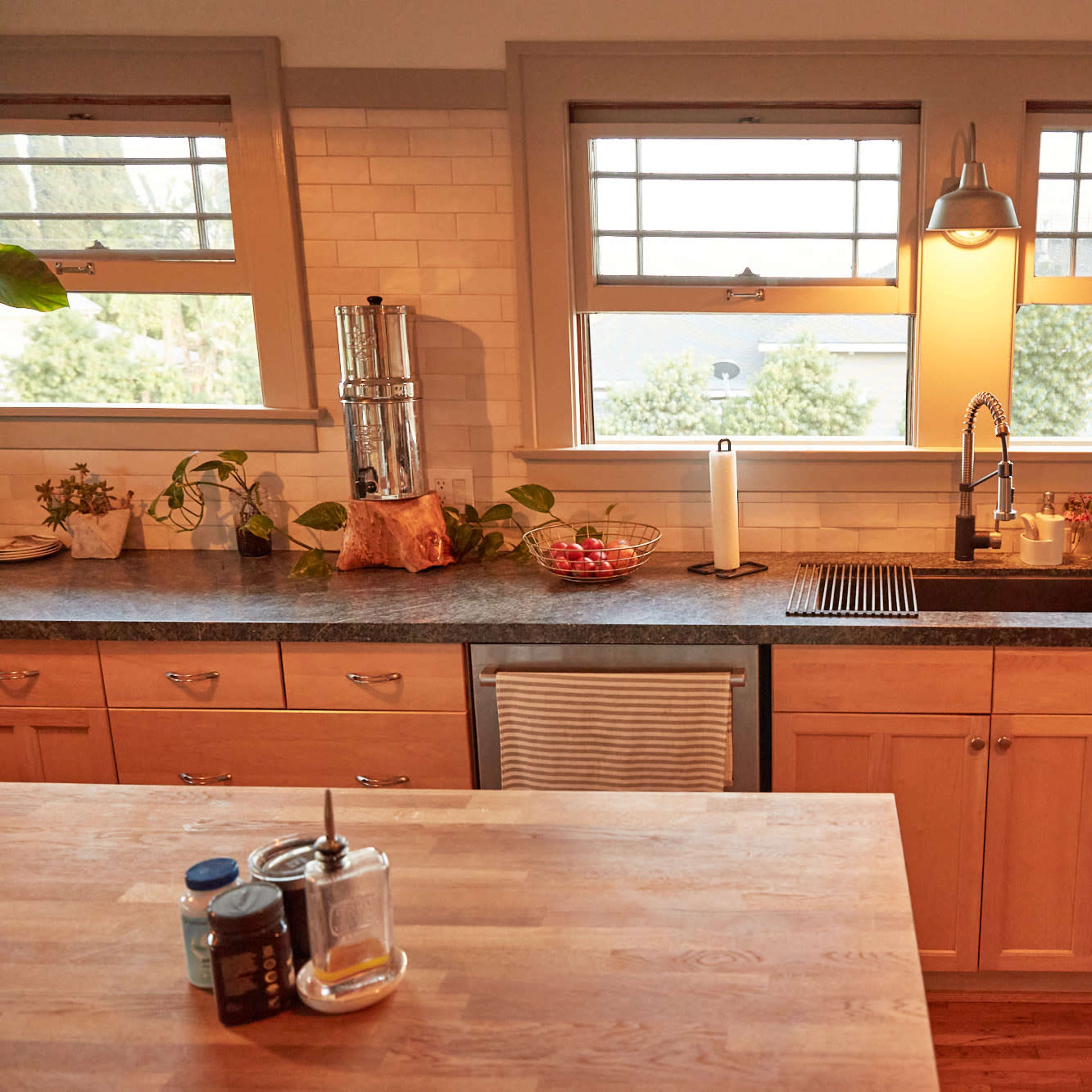 The kitchen features wood cabinetry, a stainless steel sink, and a countertop with various kitchen items, illuminated by natural light from nearby windows.