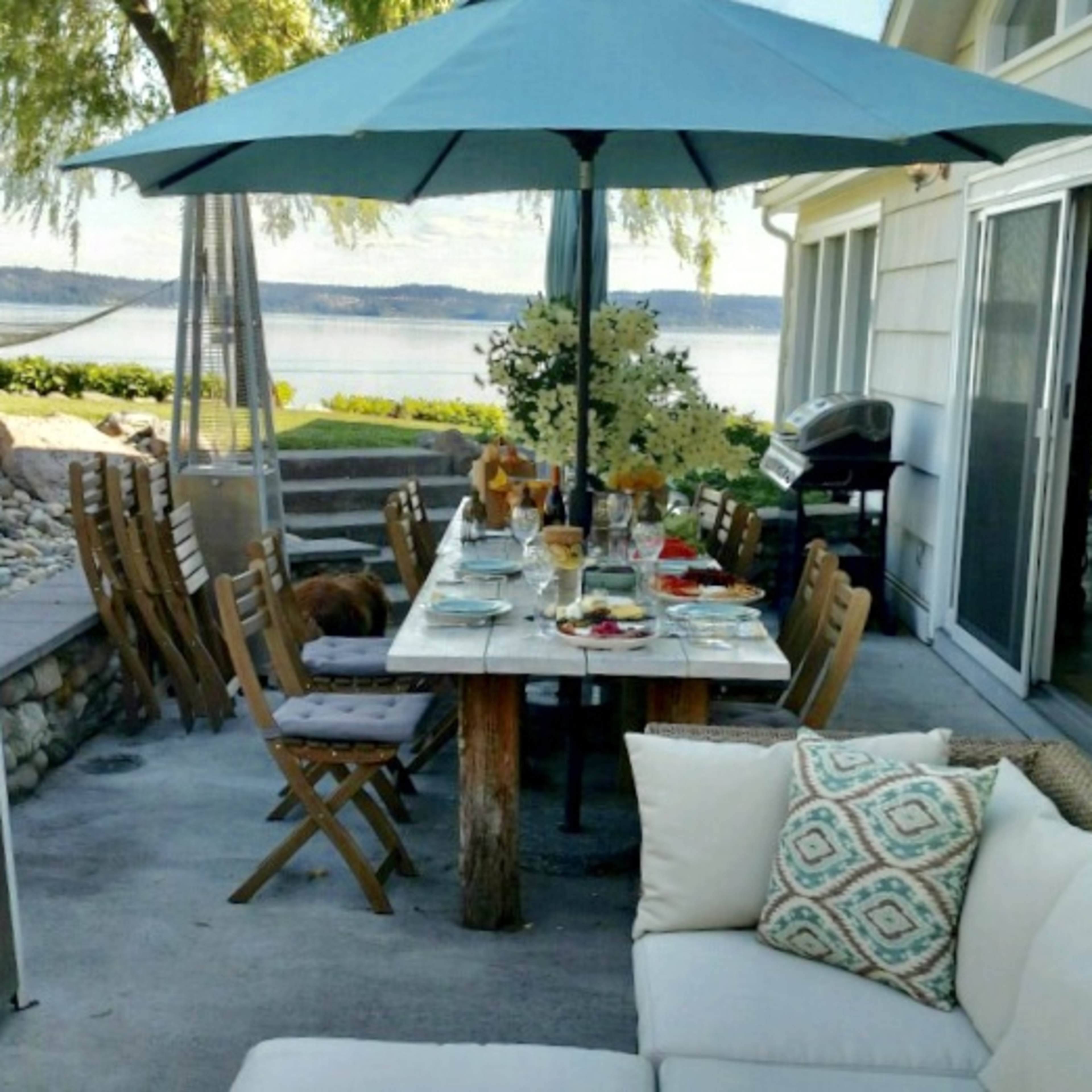 A dining table is set with food and drinks under an umbrella on a patio overlooking a lake.
