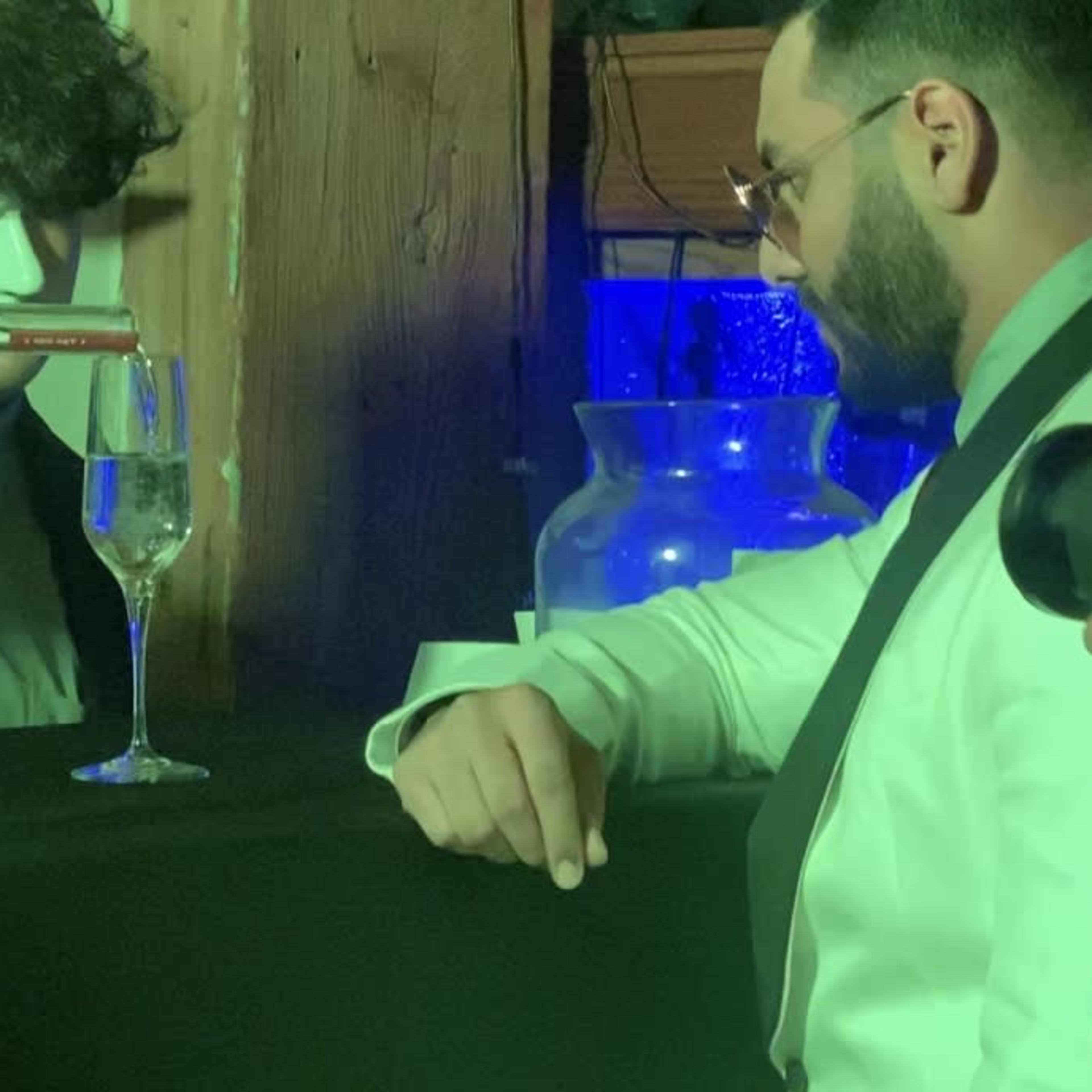 A bartender pours a drink into a glass while a man in a white suit observes at a bar counter filled with various bottles.