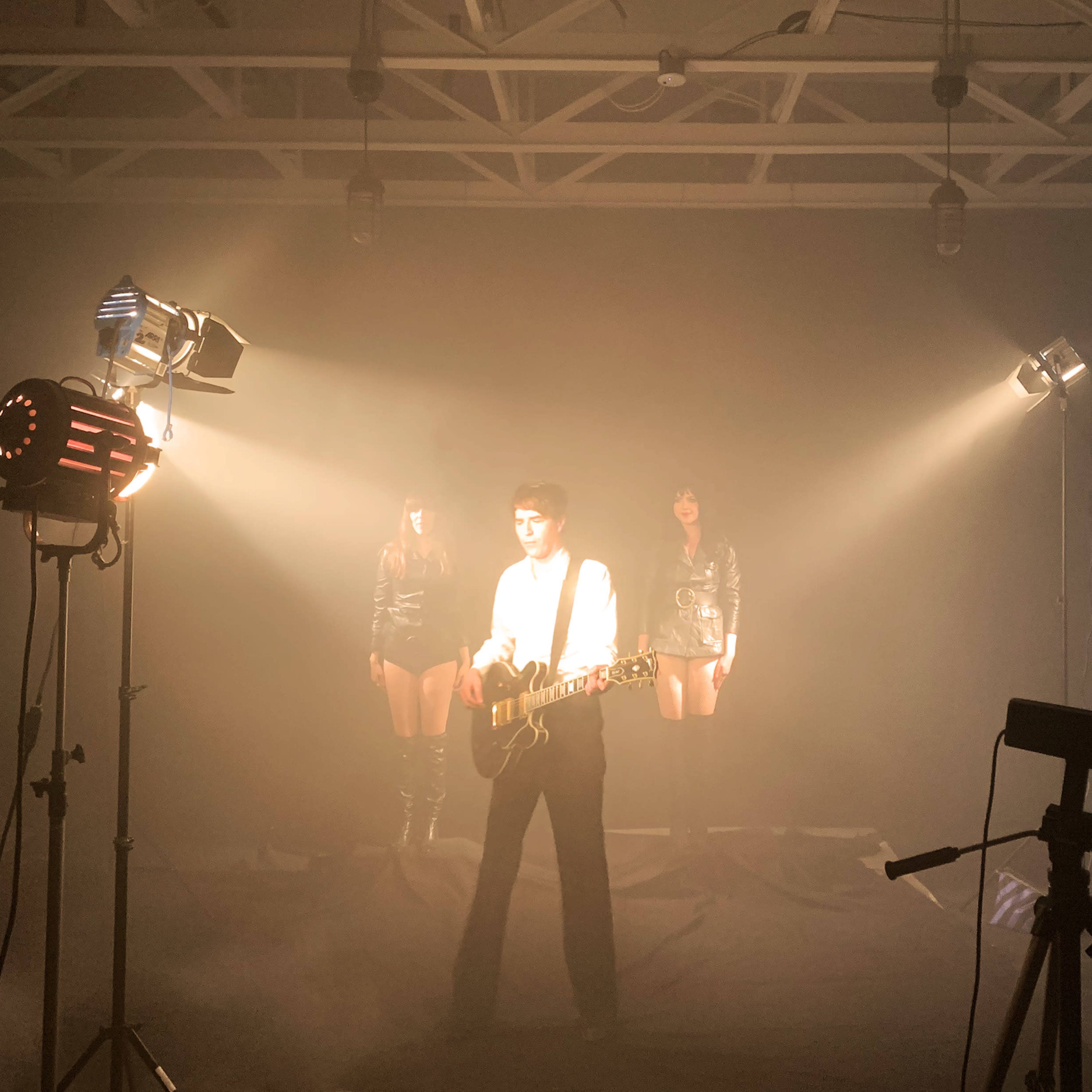 A musician plays guitar in a dimly lit space with two figures in the background, flanked by spotlight beams.