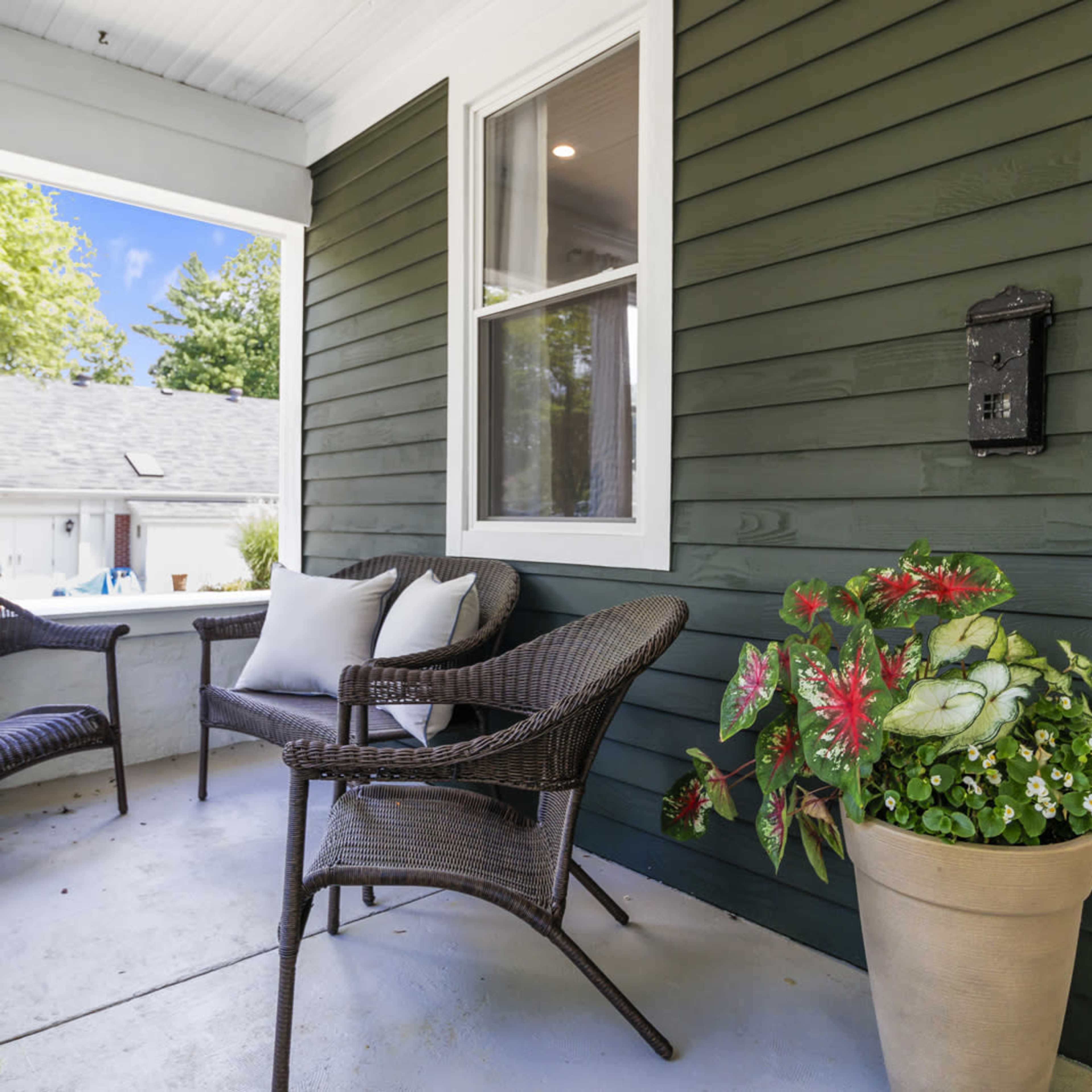 A front porch with two wicker chairs, a small table, and a potted plant next to the entrance of a house with green siding.