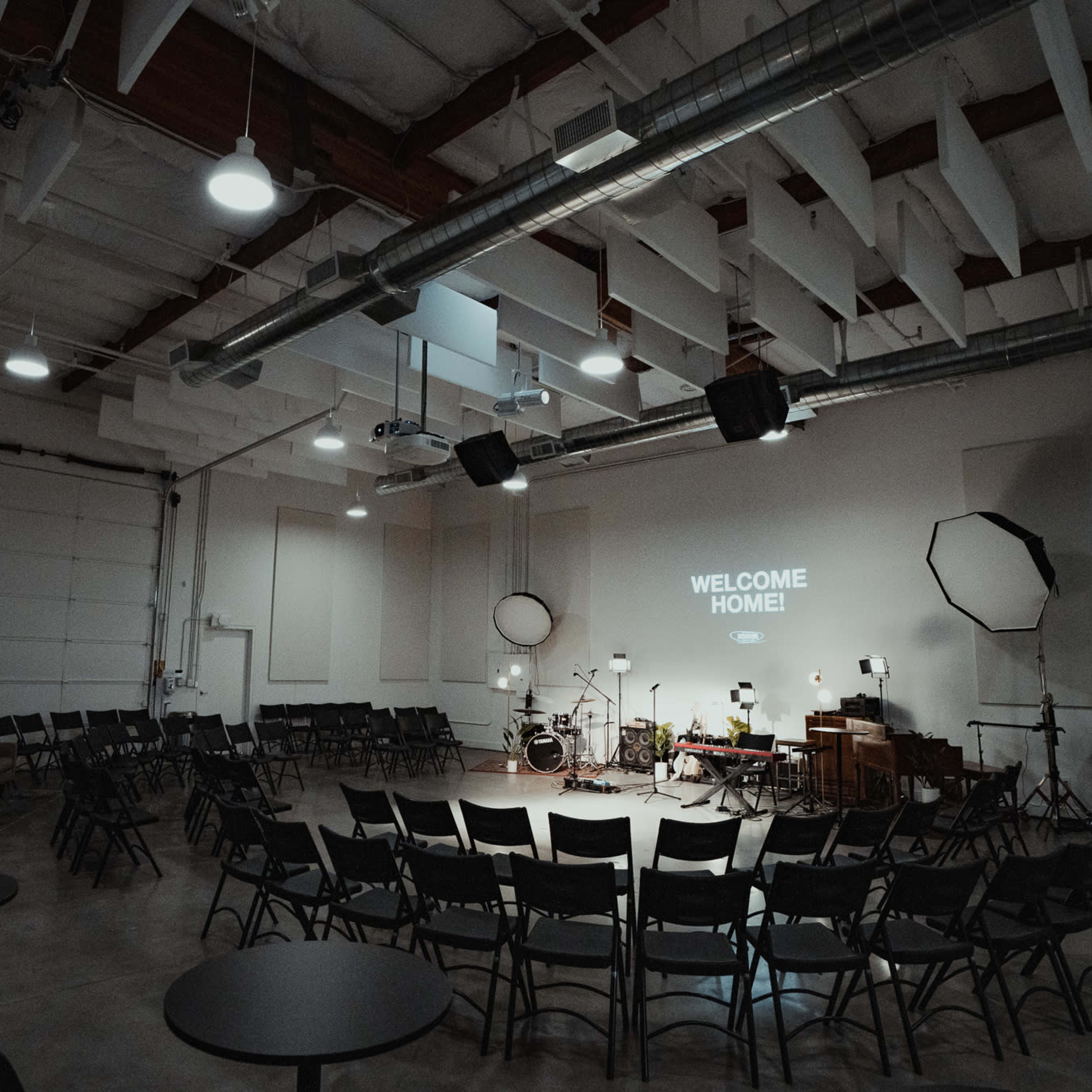 A spacious room is set up with circular rows of chairs facing a stage featuring a projector screen that reads "WELCOME HOME!" alongside musical instruments and lighting equipment.