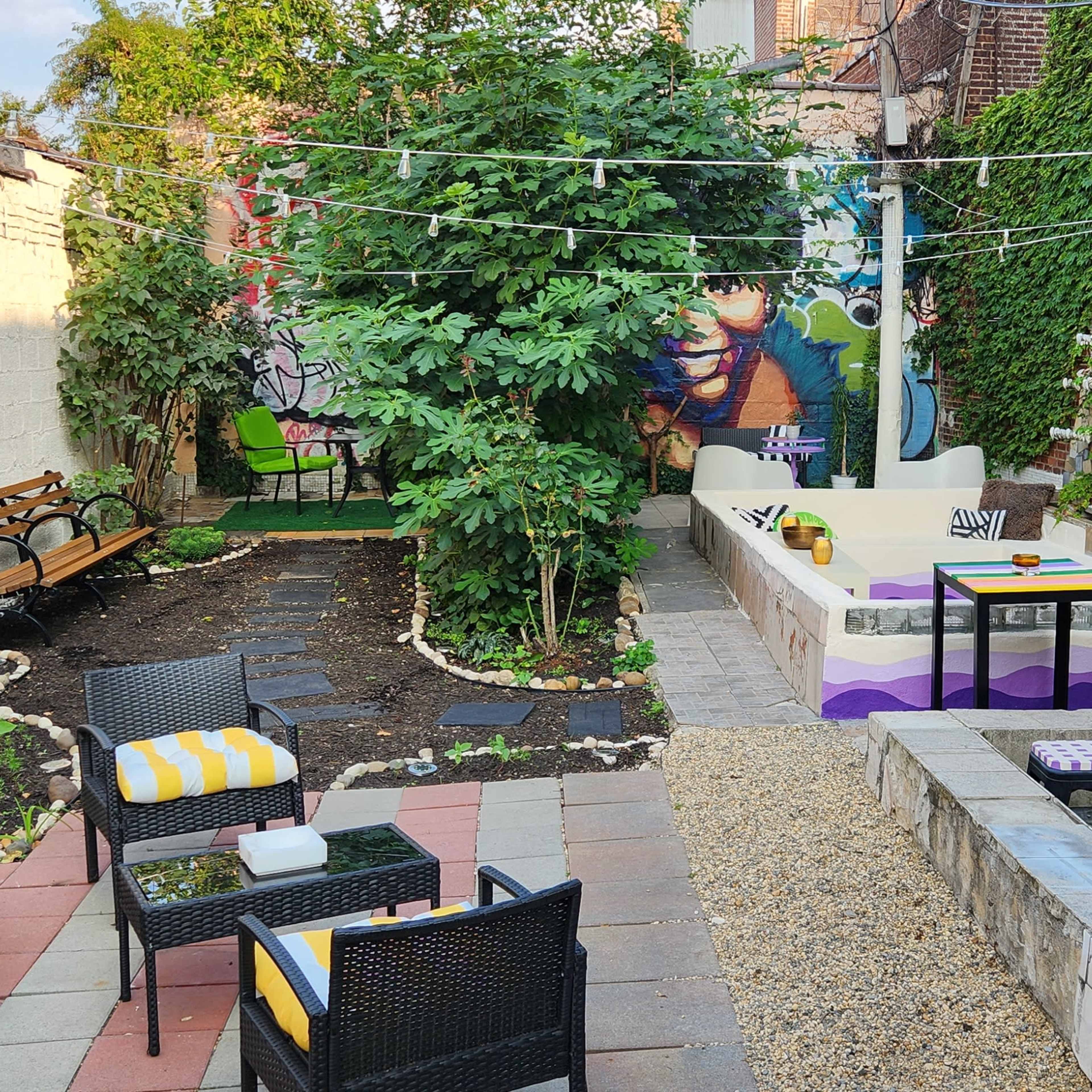 A landscaped backyard with seating areas, a fire pit, and a mural-covered wall, surrounded by greenery.