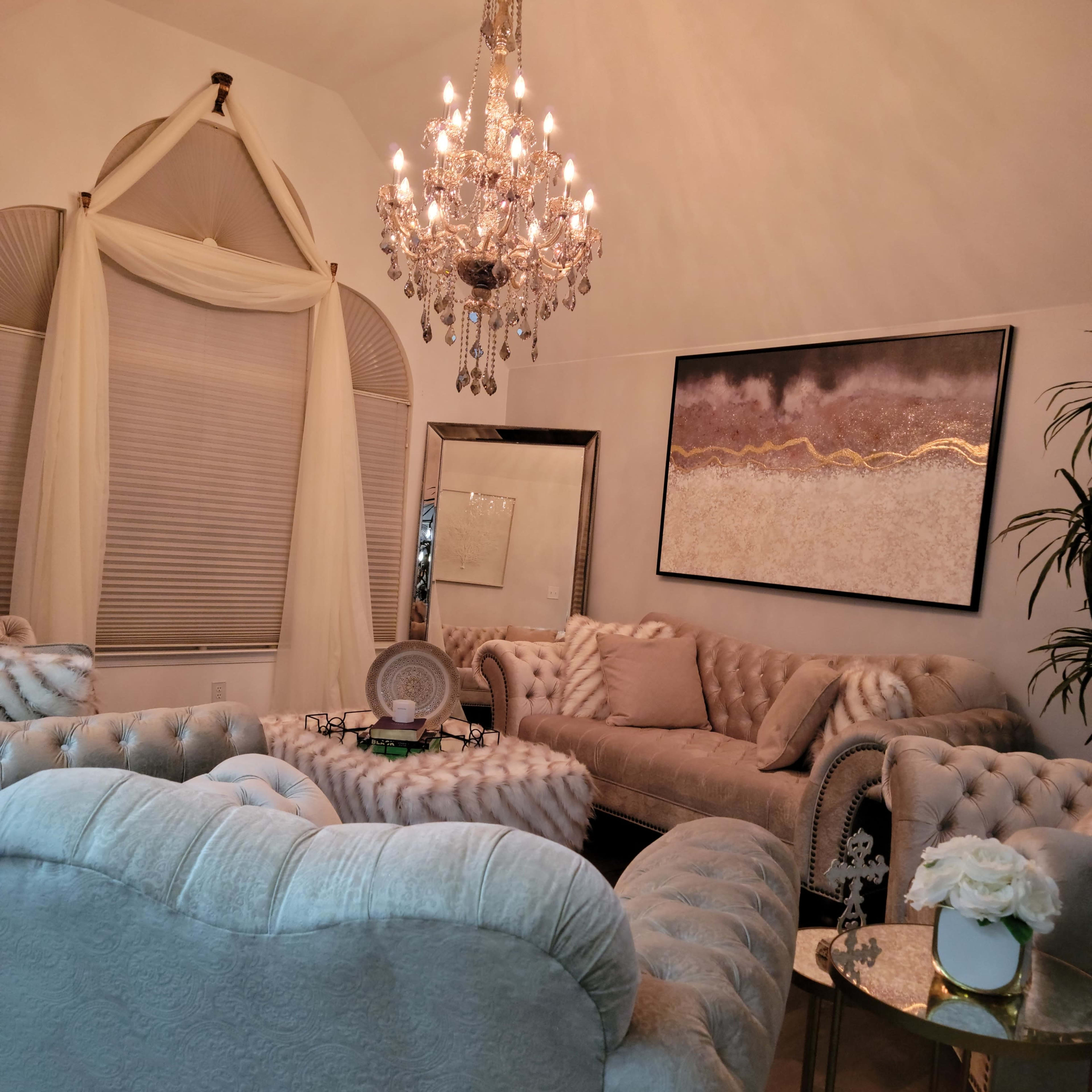 A elegantly furnished living room featuring plush sofas, a chandelier, and decorative elements including a mirror and a small table with floral arrangements.