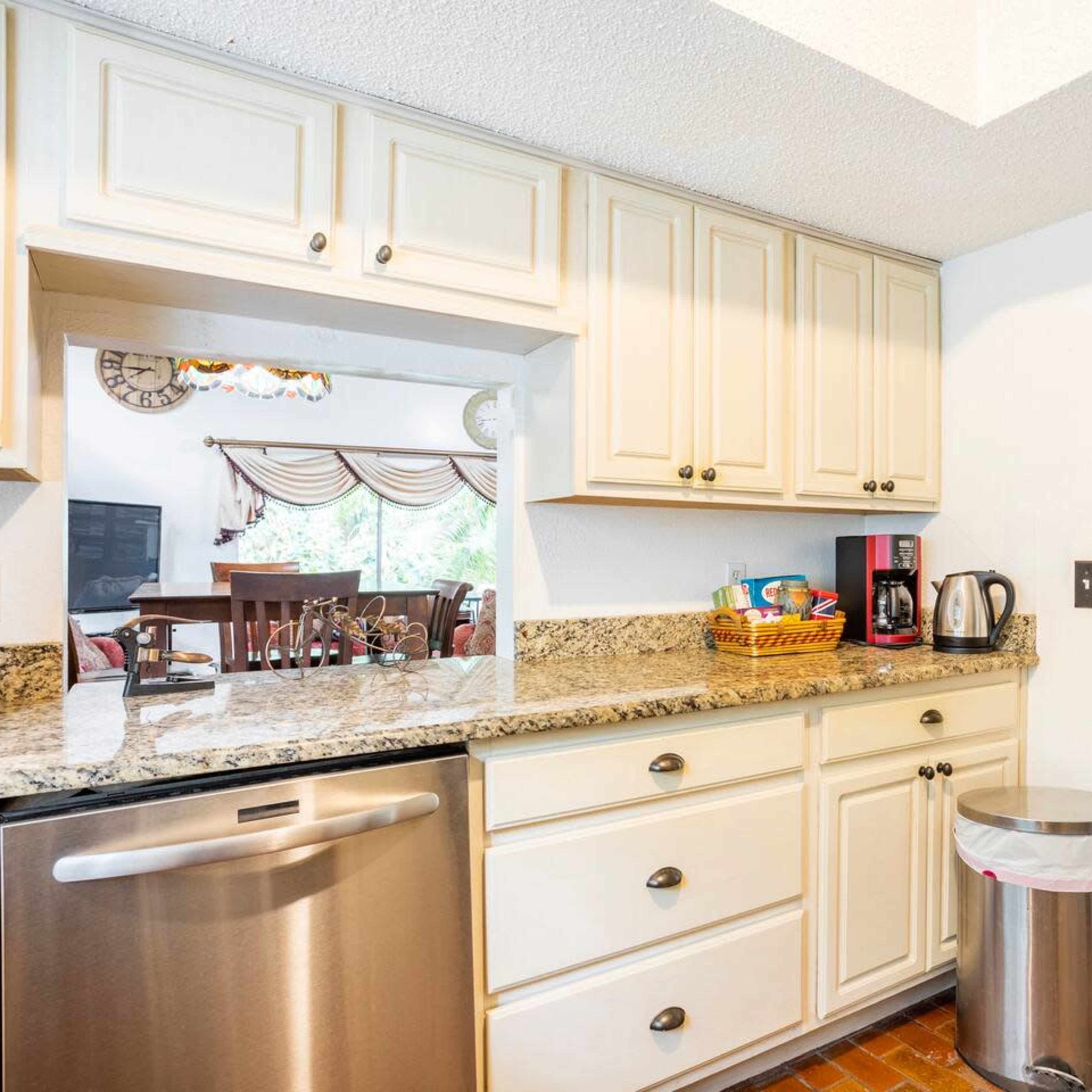 A kitchen features cream-colored cabinets, a granite countertop, stainless steel appliances, and an open view to a dining area and bathroom beyond.