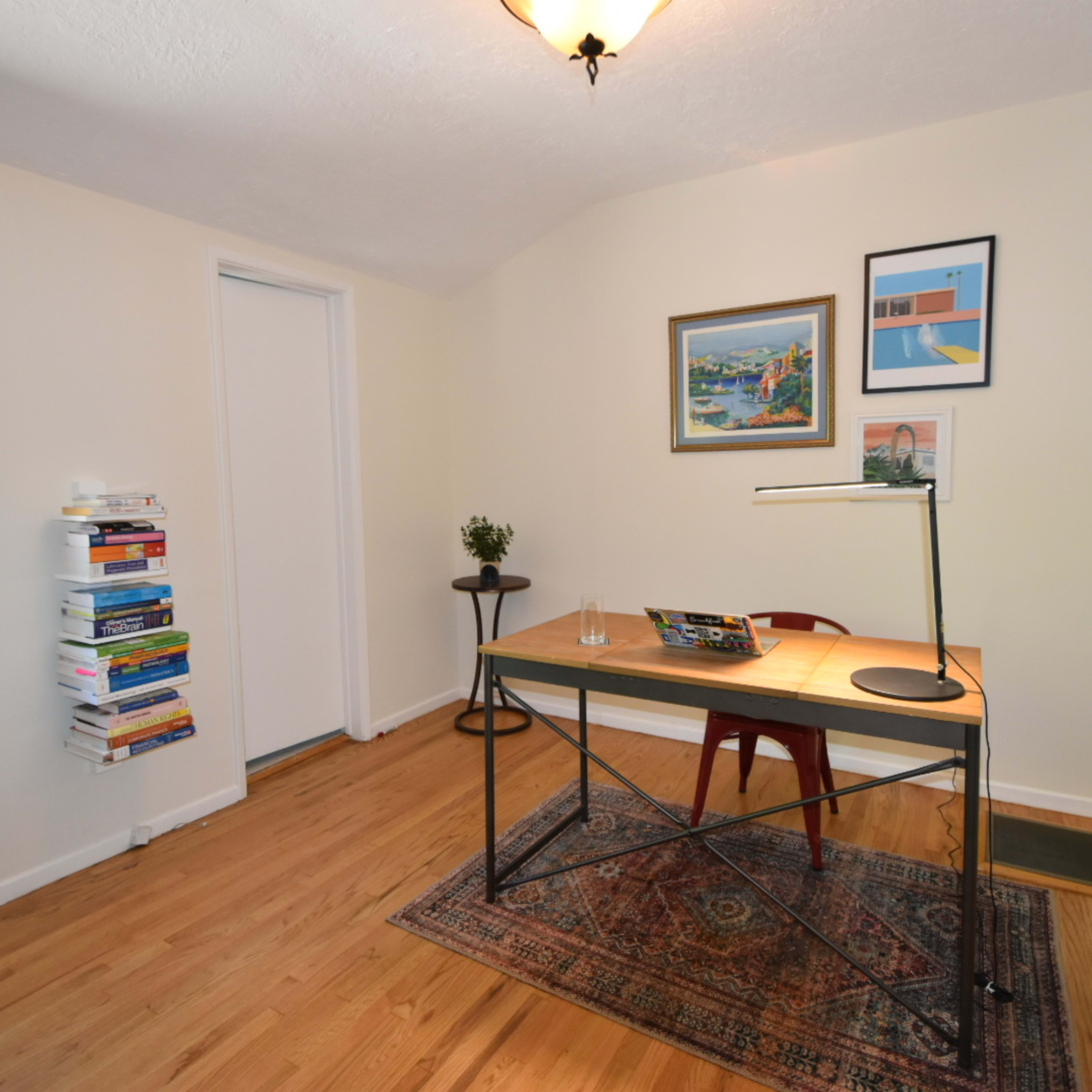 A minimalist home office with a wooden desk, a chair, a lamp, and a small bookshelf stacked with books.