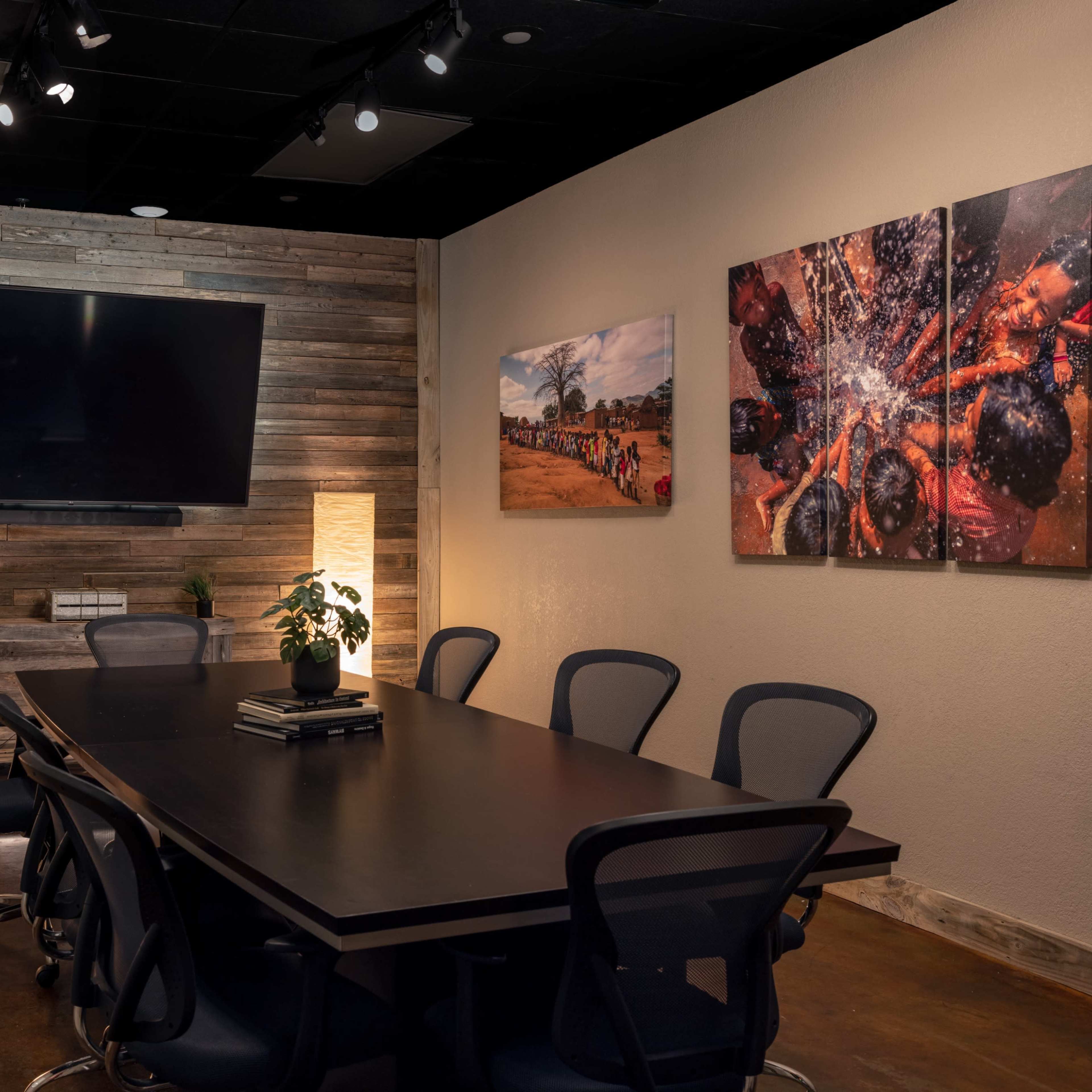A modern conference room featuring a large table surrounded by chairs, a wall-mounted television, and artwork on the walls illuminated by soft floor lamps.