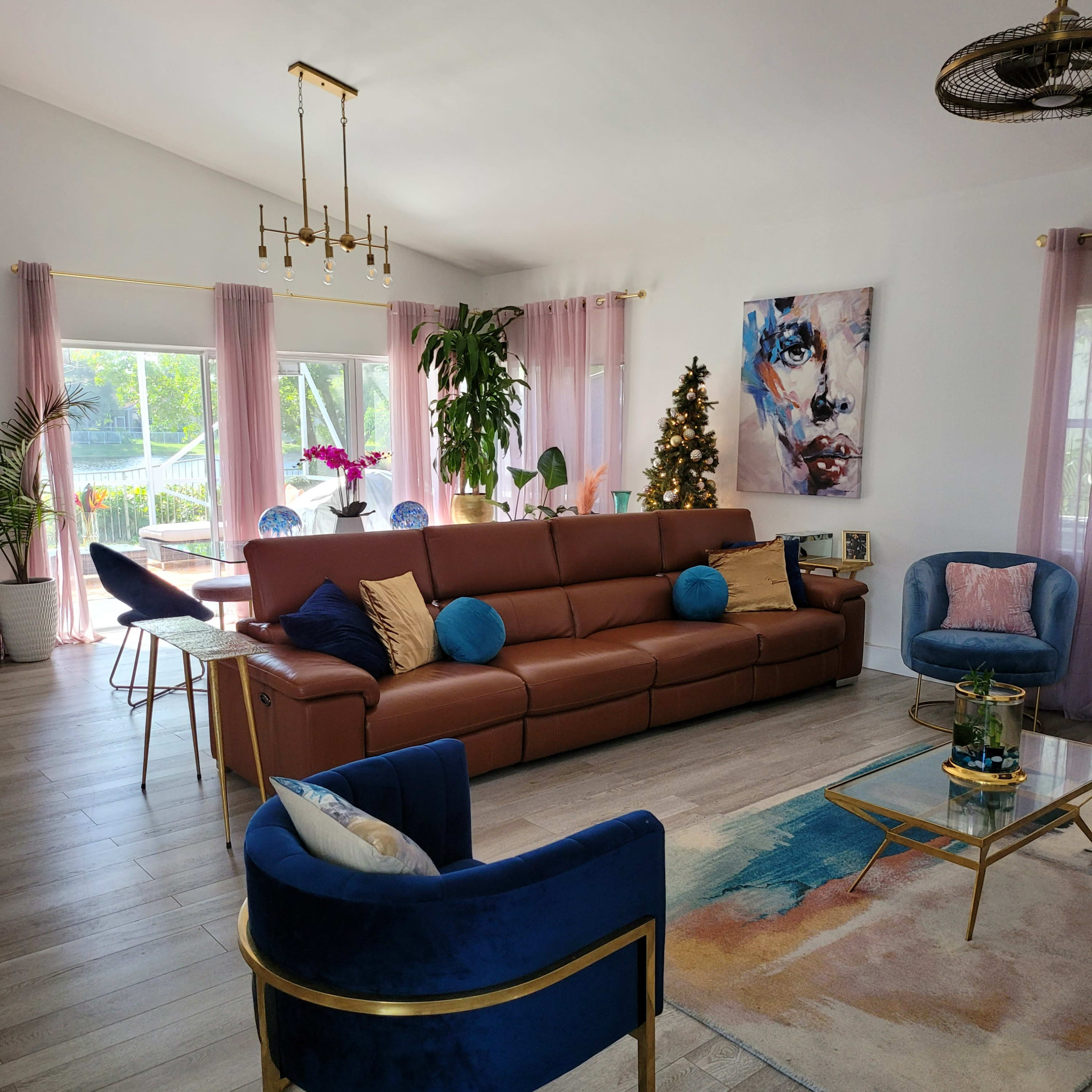 A modern living room with a brown sectional sofa, two blue accent chairs, a glass coffee table, and large windows displaying greenery outside.