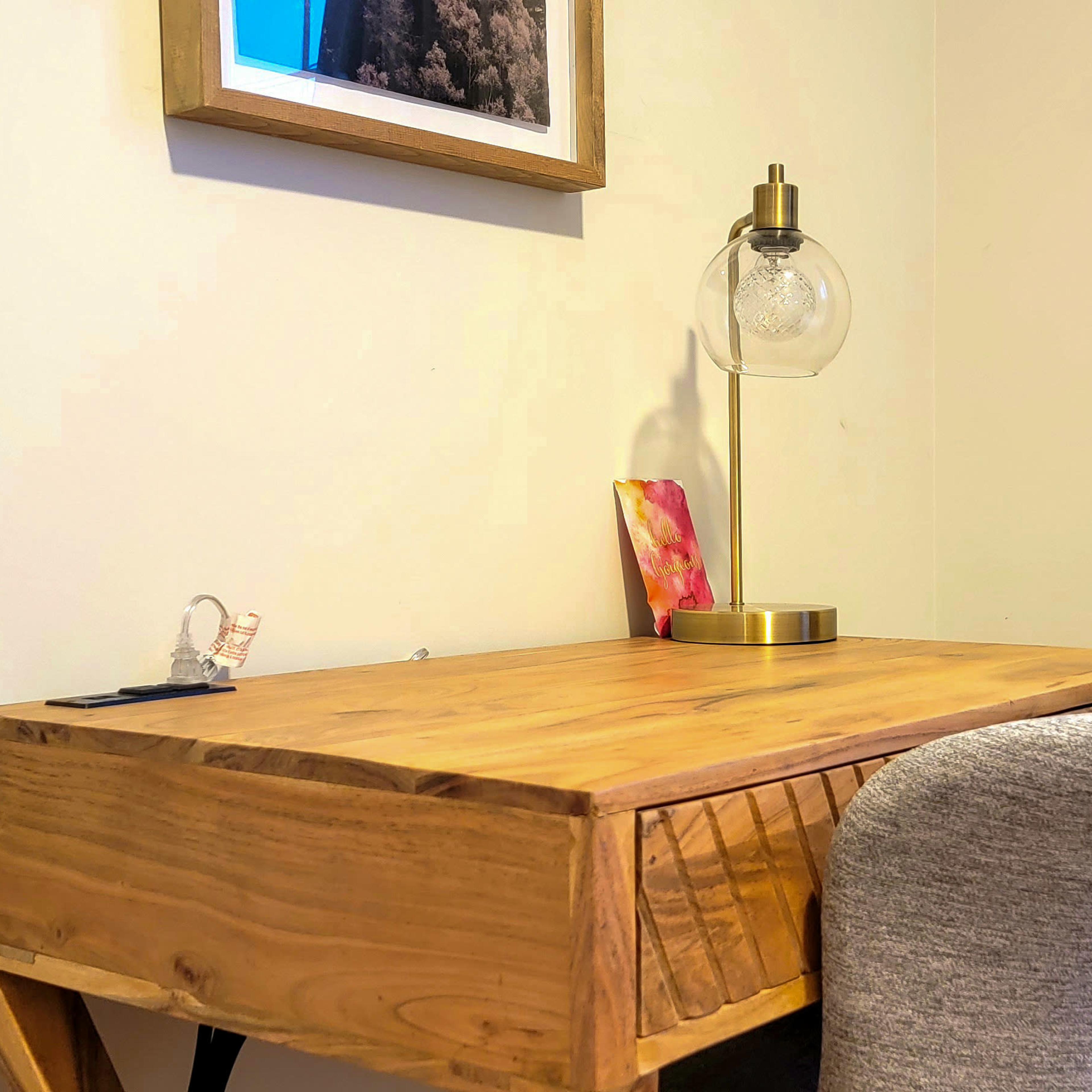 A wooden desk with a drawer, a decorative lamp, and a framed picture is positioned against a wall beside a curtain.
