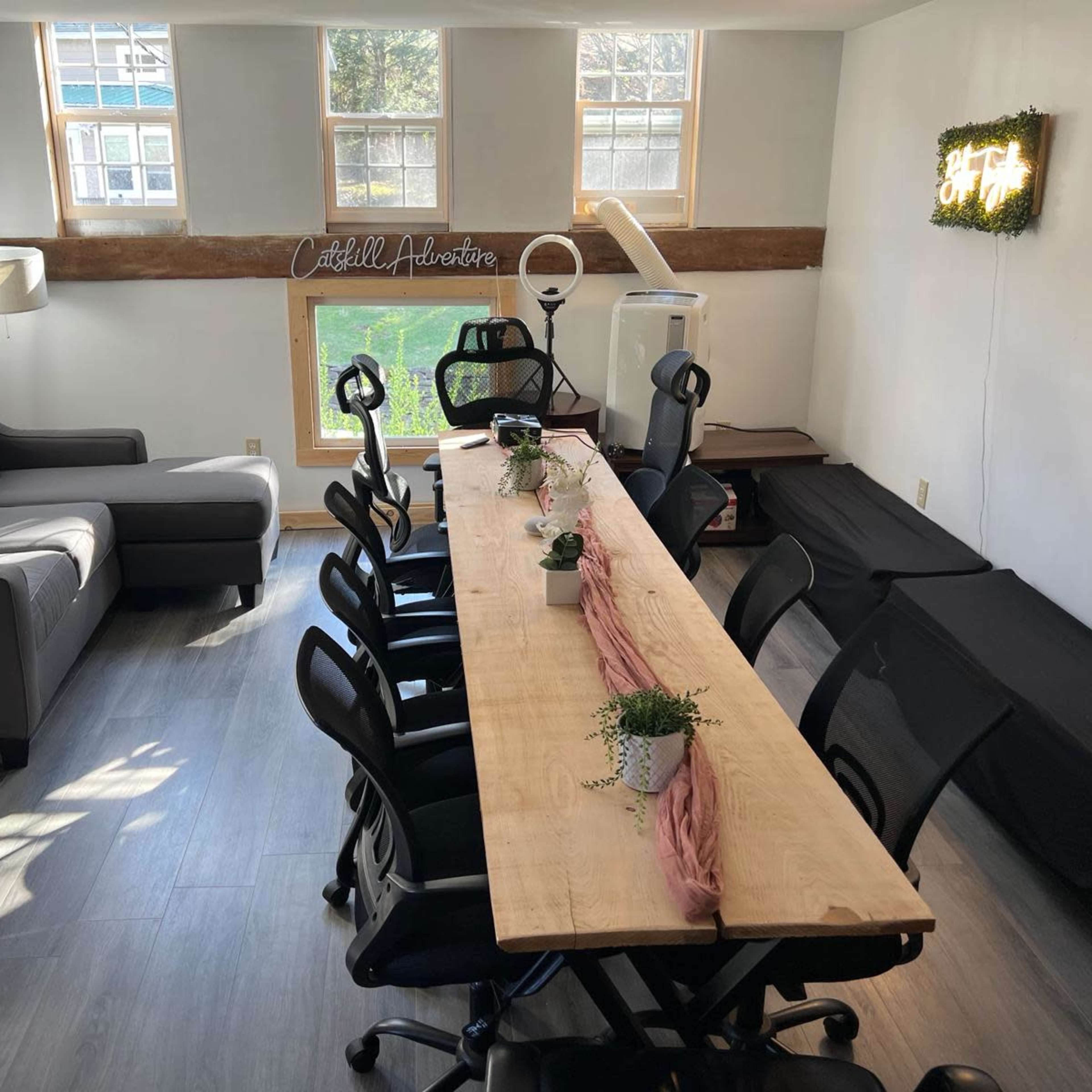 A well-lit meeting room with a long wooden table surrounded by black office chairs, a sofa in the corner, and a window that offers a view of greenery outside.
