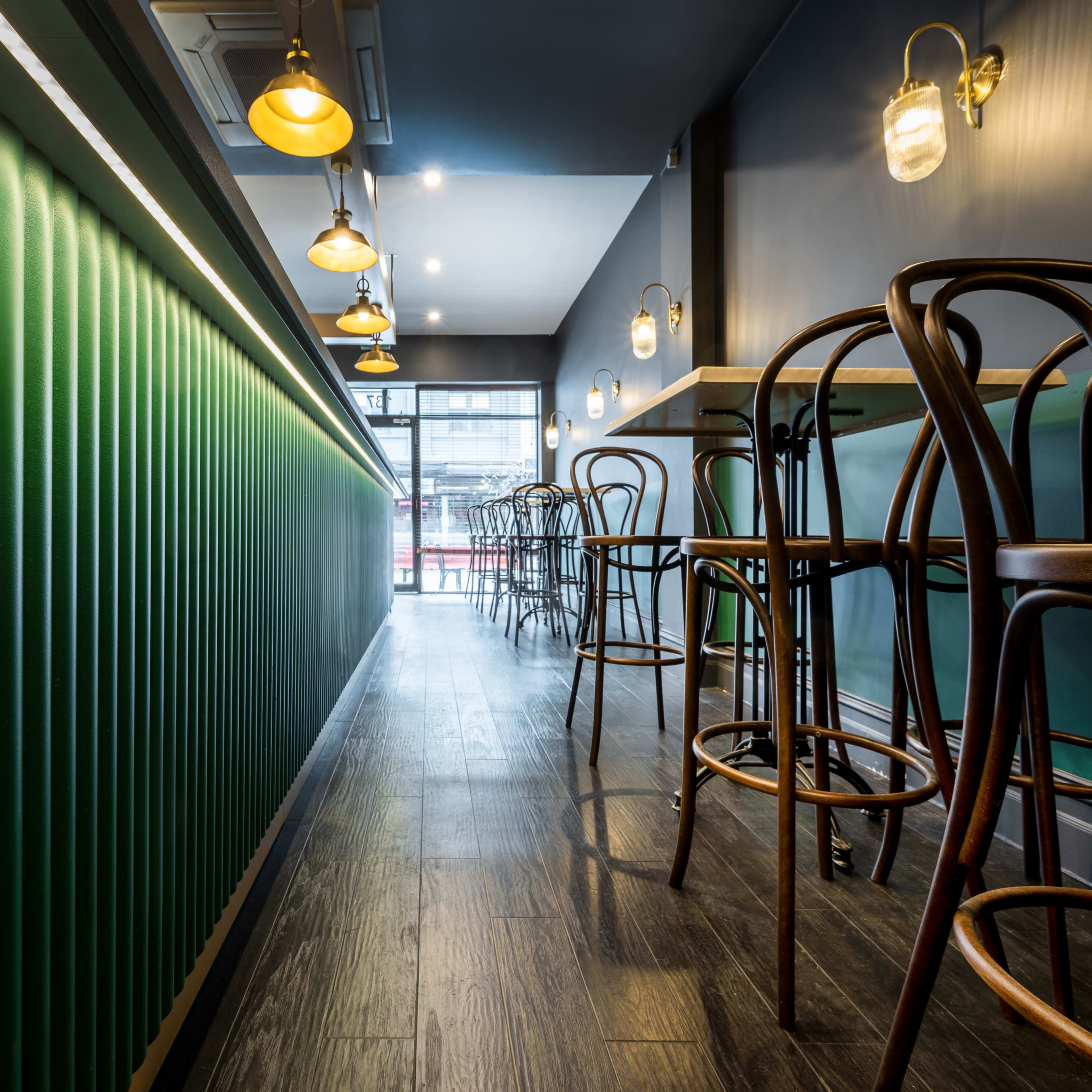 A modern café interior featuring green paneled walls, wooden flooring, and high stools arranged along a counter with pendant lighting.
