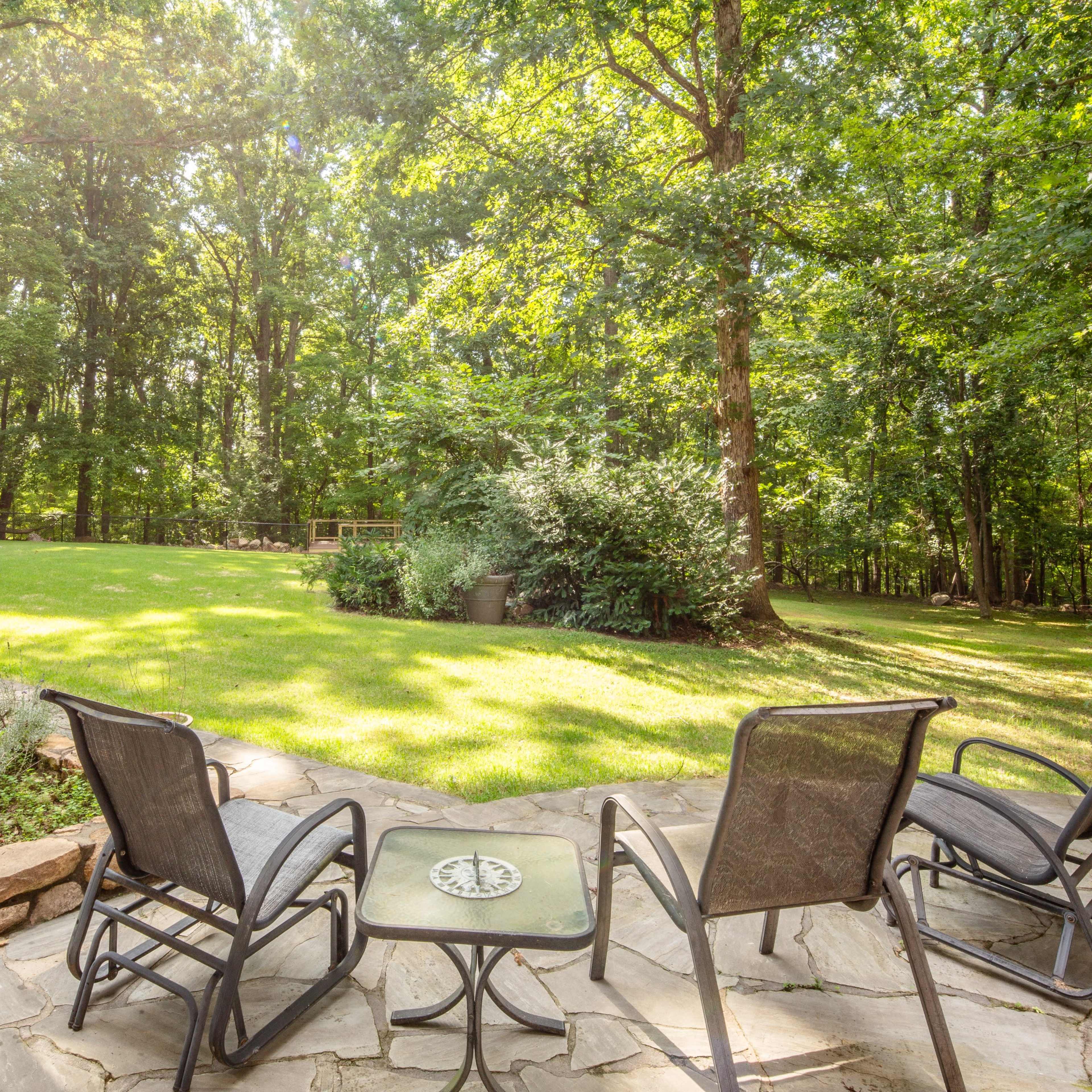 A stone patio with four chairs and a table overlooks a grassy yard surrounded by trees.