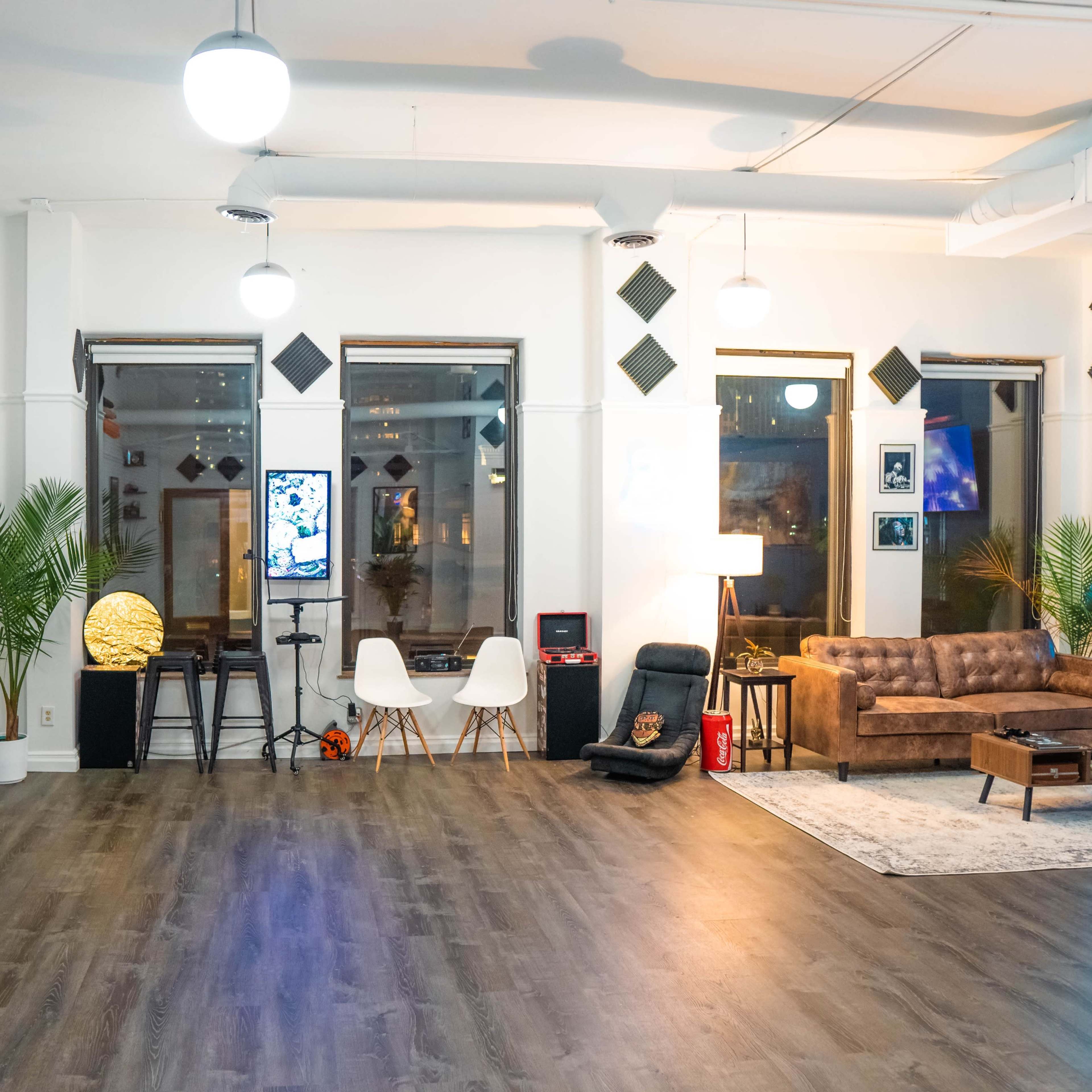 A spacious room with wooden flooring, featuring a brown leather sofa, a small table, and decorative plants alongside a large window displaying city lights.