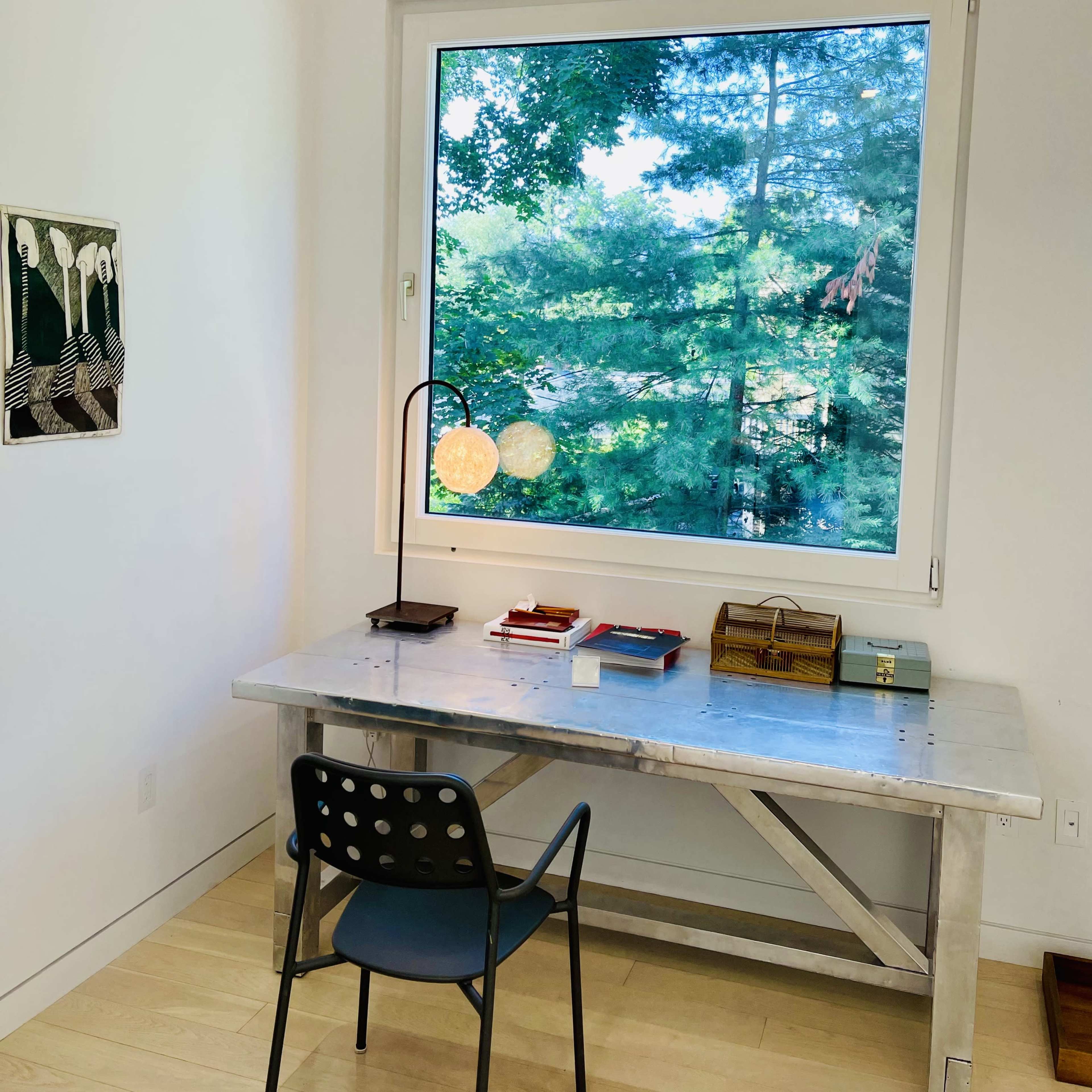 A minimalist workspace features a metal desk, a chair, and a lamp, with a large window displaying a view of trees outside.