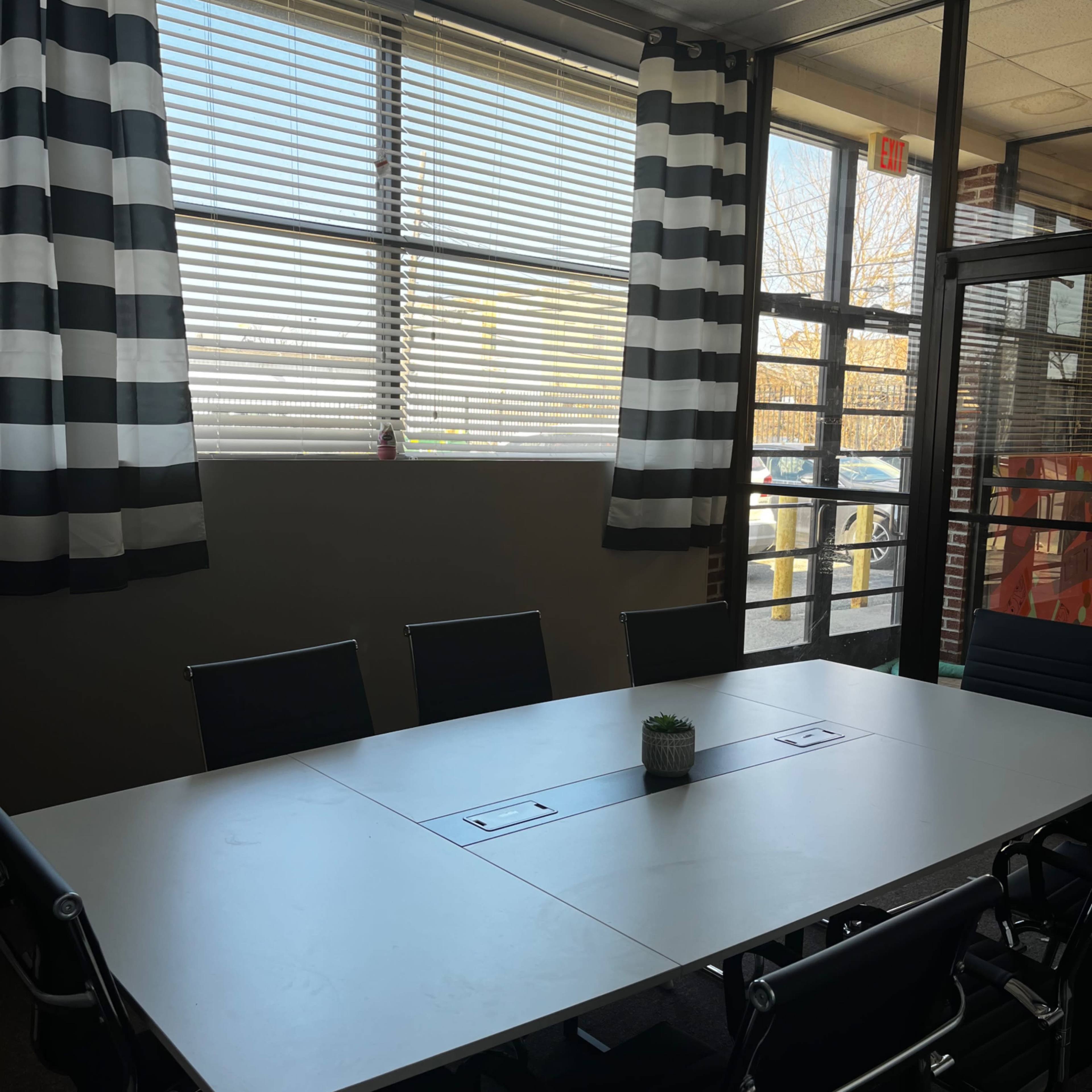 A conference room with a large table surrounded by black chairs, sunlight streaming in through large windows with striped curtains.
