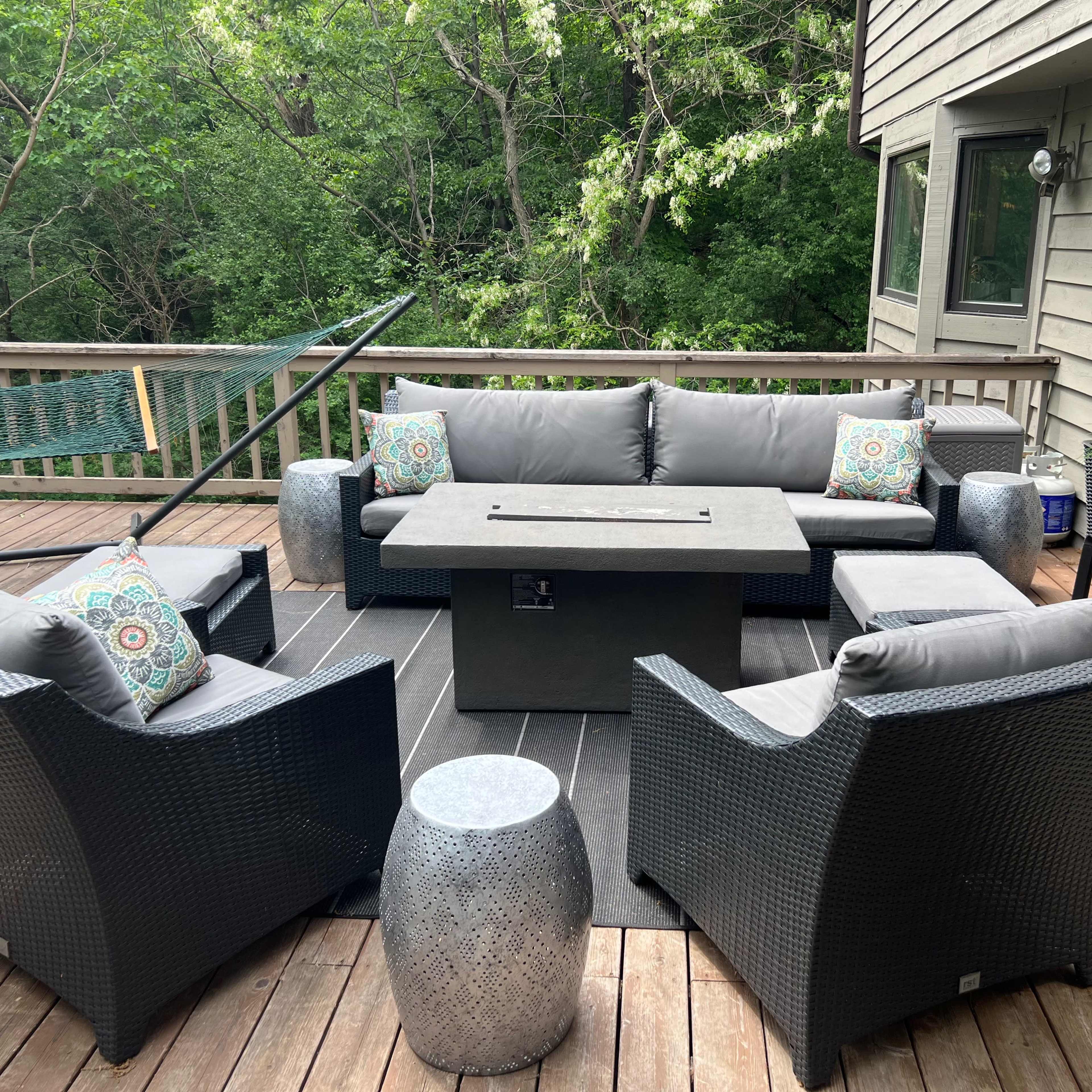 A deck features a seating area with gray outdoor furniture arranged around a central fire table, surrounded by lush greenery.
