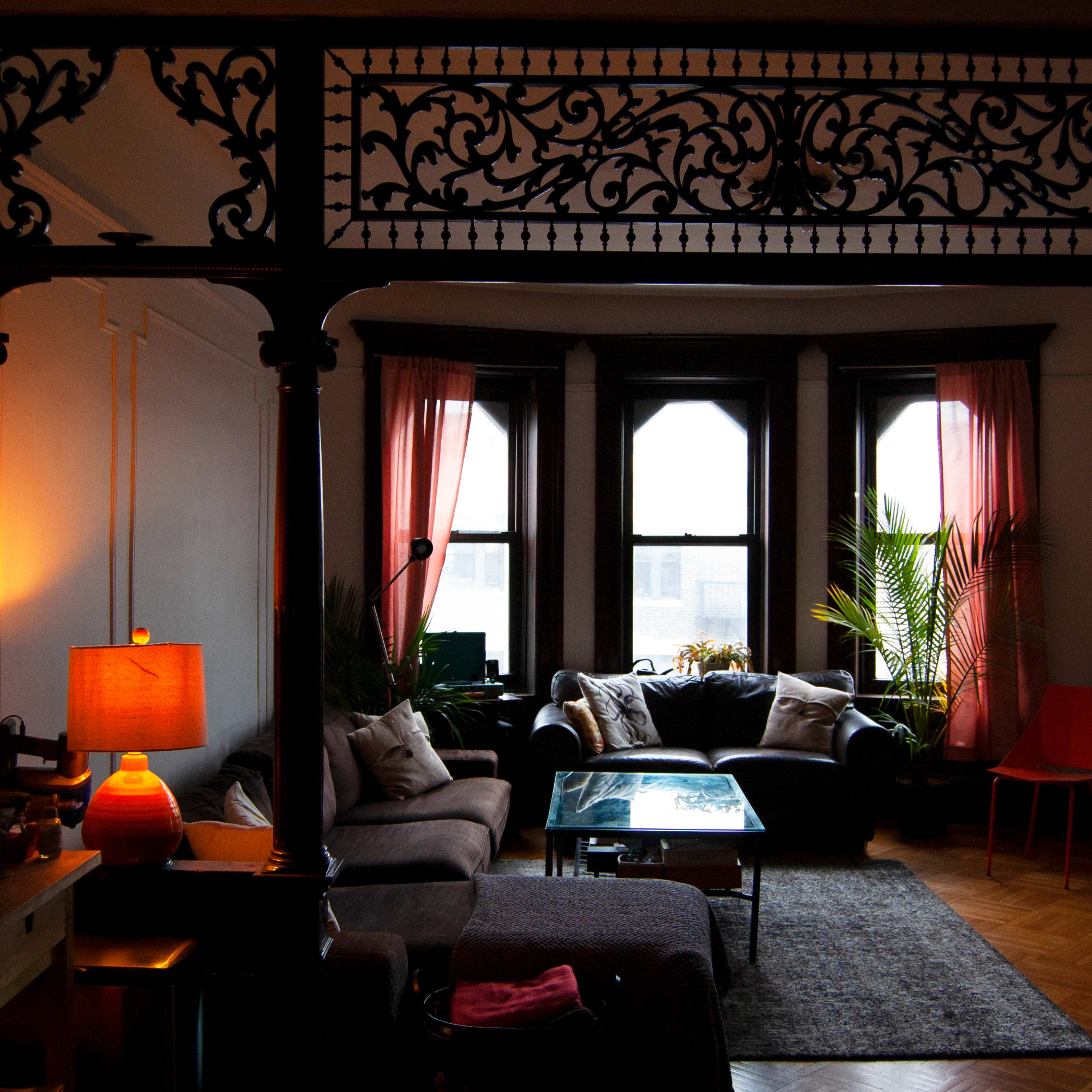 The scene features a living room with large windows draped in pink curtains, a sectional sofa, a glass coffee table, and decorative plants, all framed by an ornate architectural divider.