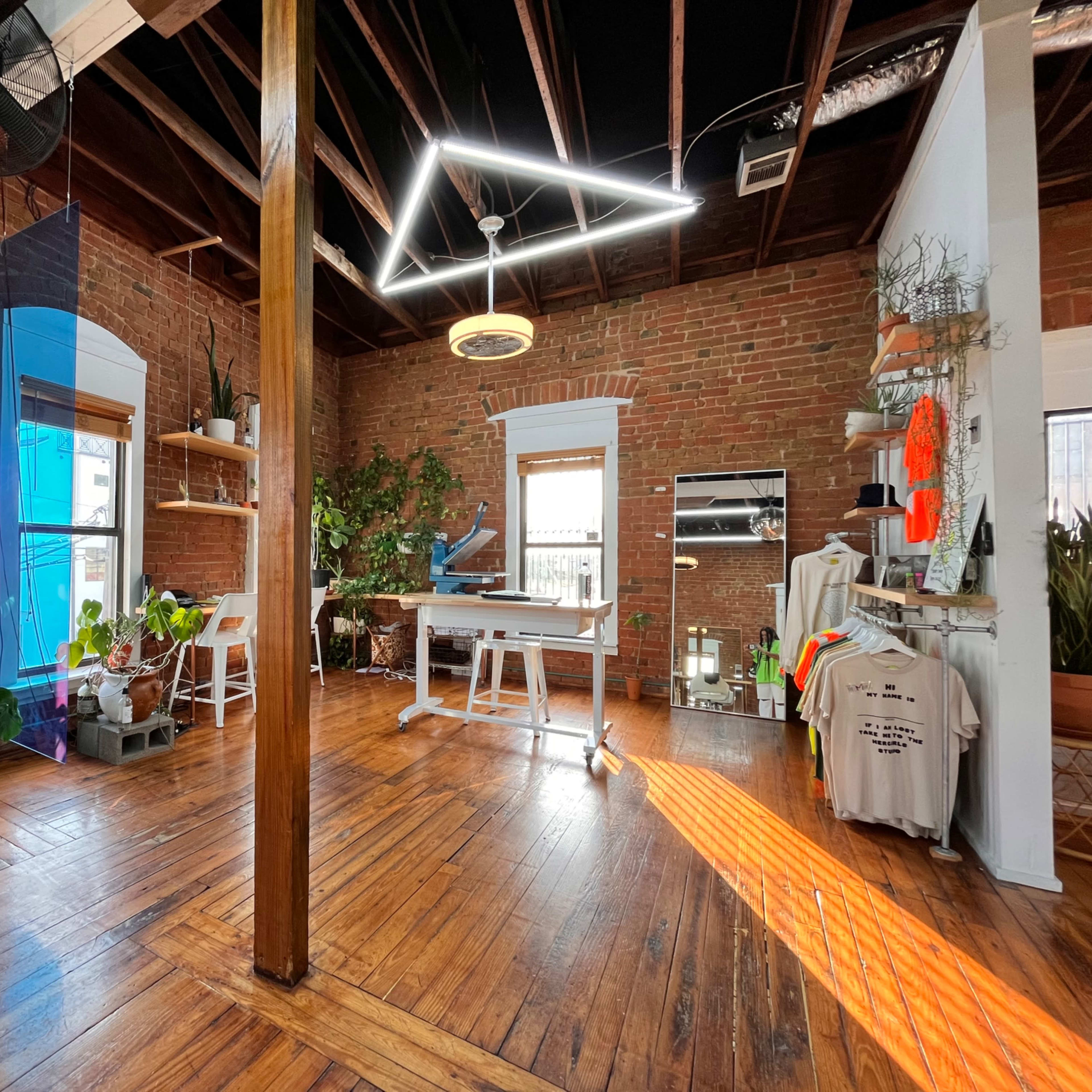 A spacious room with exposed brick walls, wooden floors, and various indoor plants, featuring a large work table, colorful decor items, and bright natural light streaming through the windows.