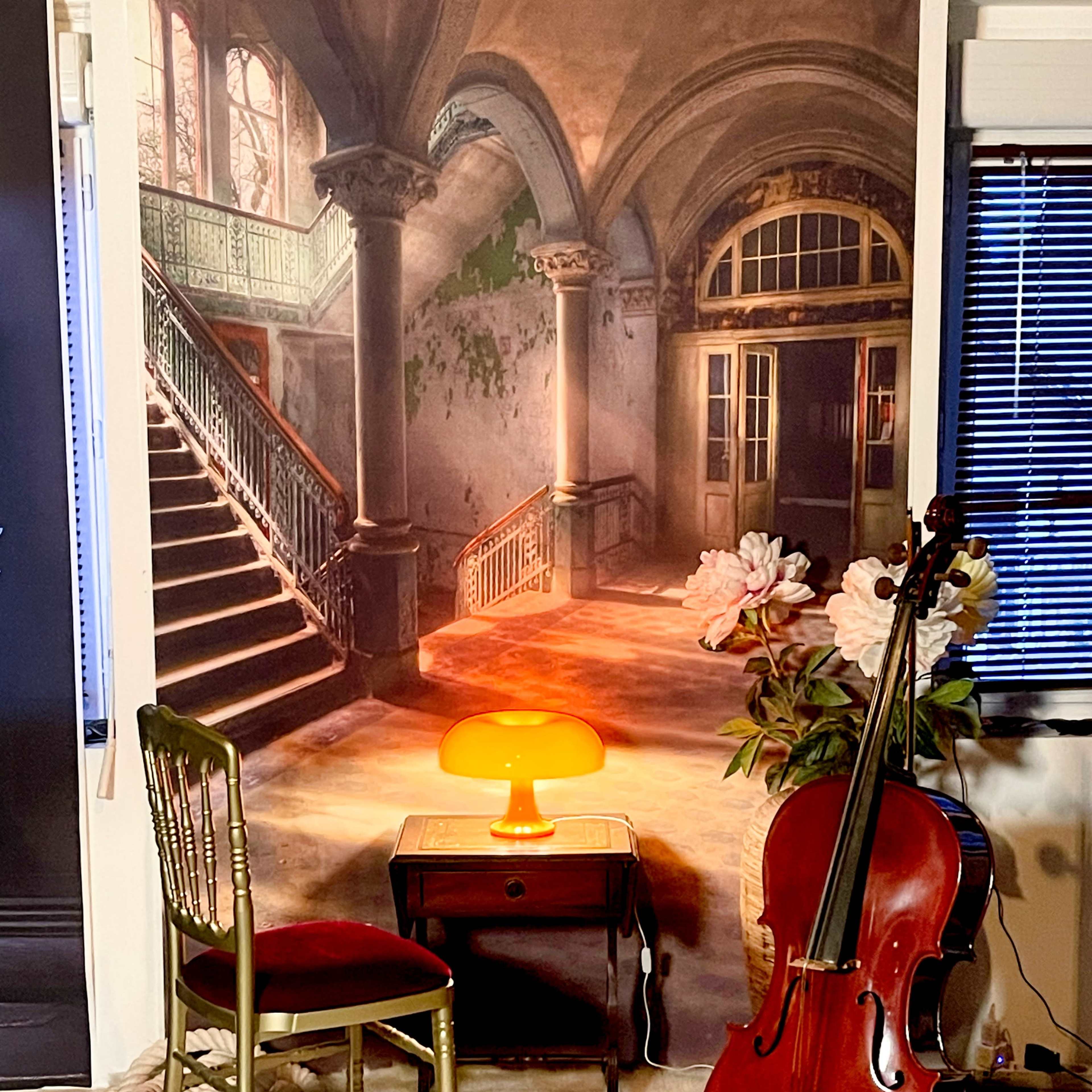 A cello stands next to a table with a lamp and a chair in front of a mural depicting a grand, dilapidated staircase and doorway.