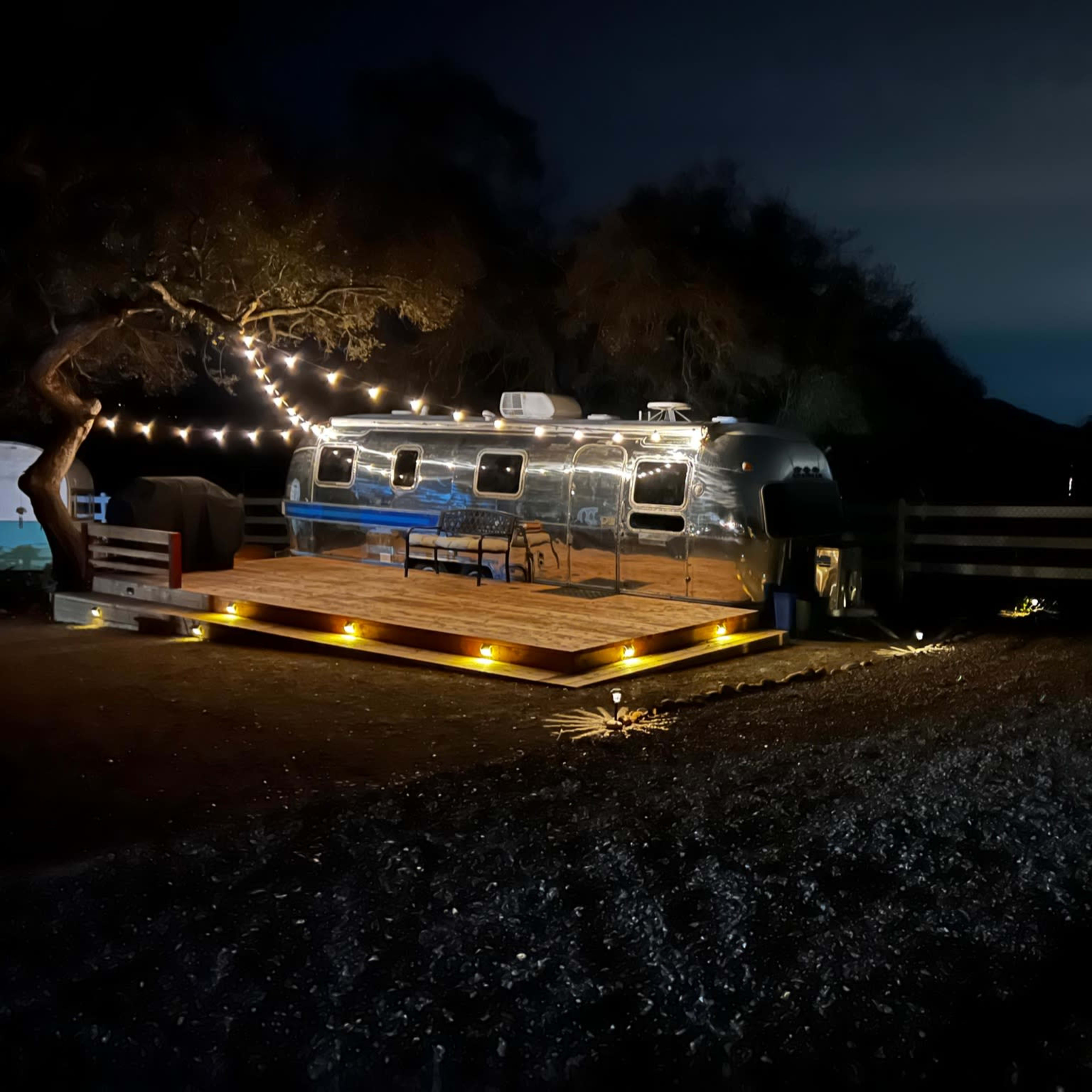 A silver Airstream trailer is parked on a wooden deck illuminated by string lights at night.
