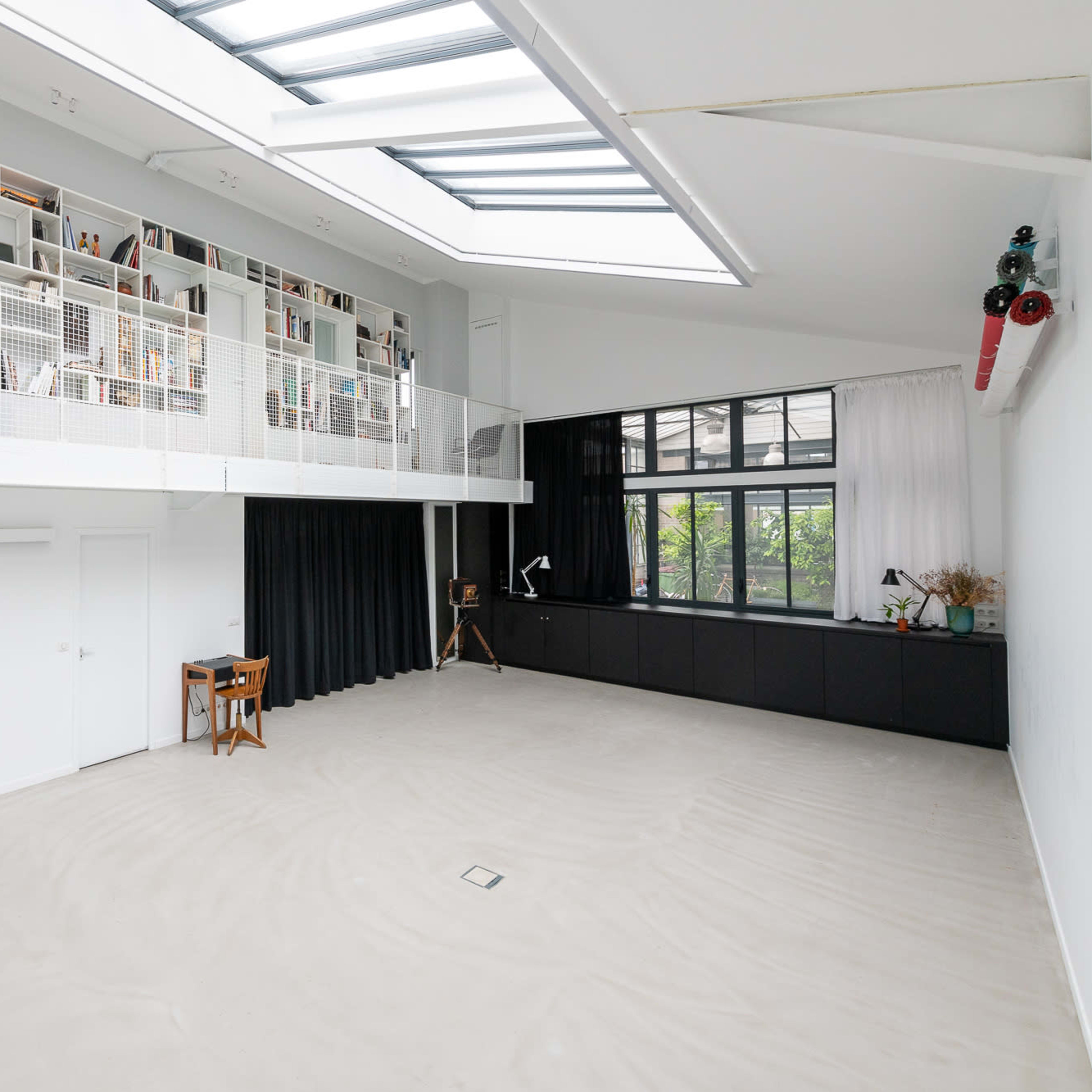 A spacious, modern interior with a high ceiling, a loft area lined with bookshelves, and large windows allowing natural light to enter.