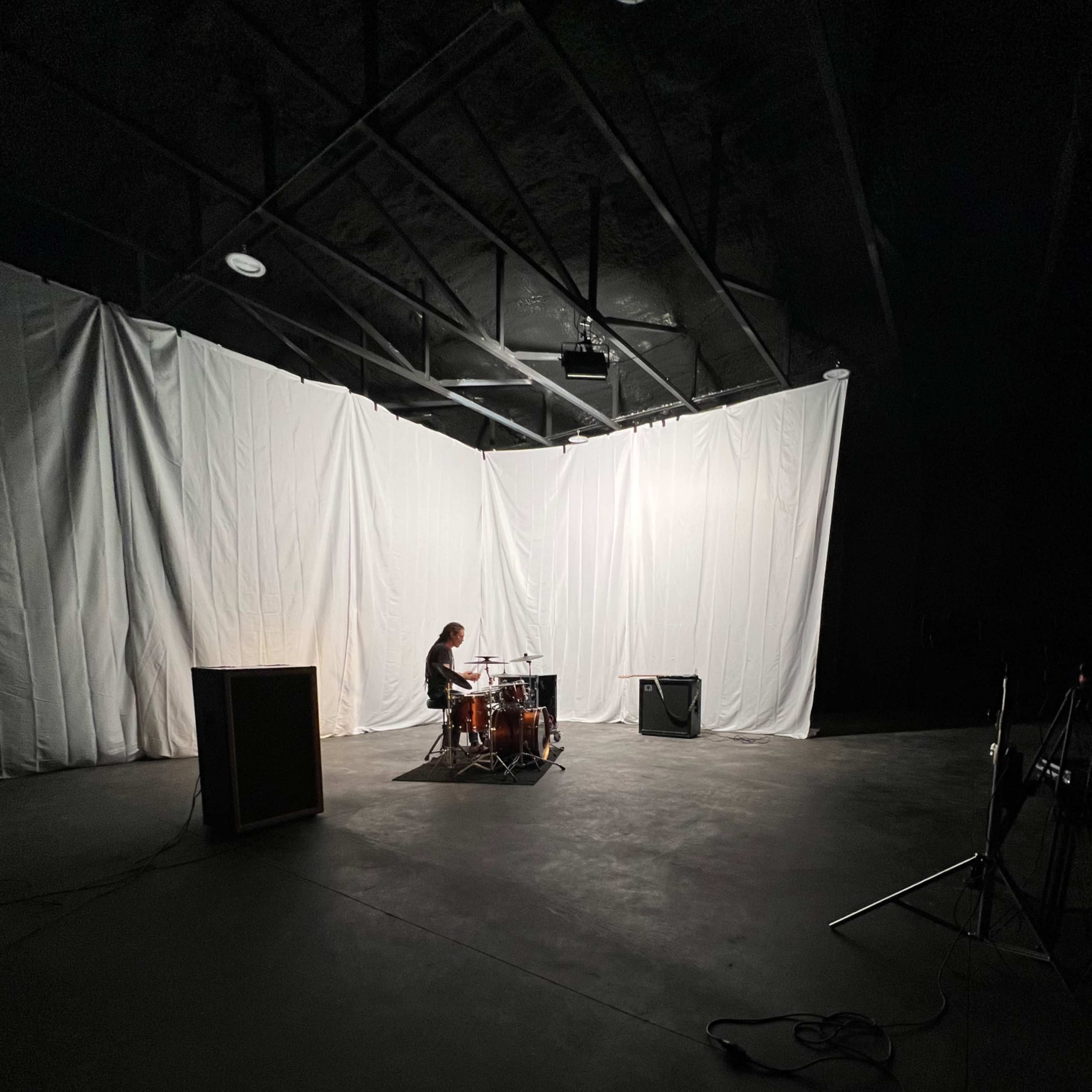 A drummer is seated at a drum set in a dimly lit space with white draping in the background.