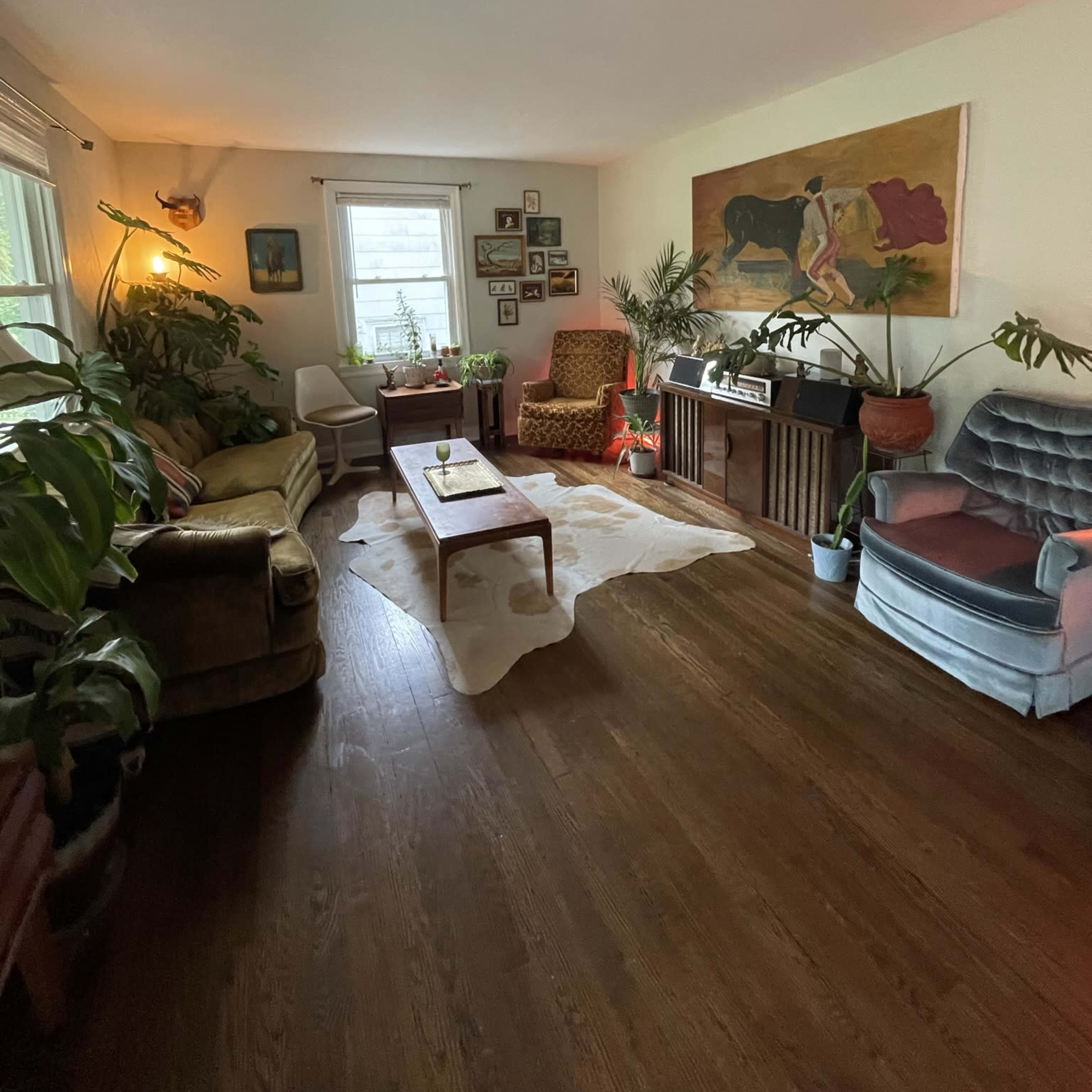A cozy living room with two patterned chairs, a sofa, a wooden coffee table, various plants, and a large artwork on one wall.