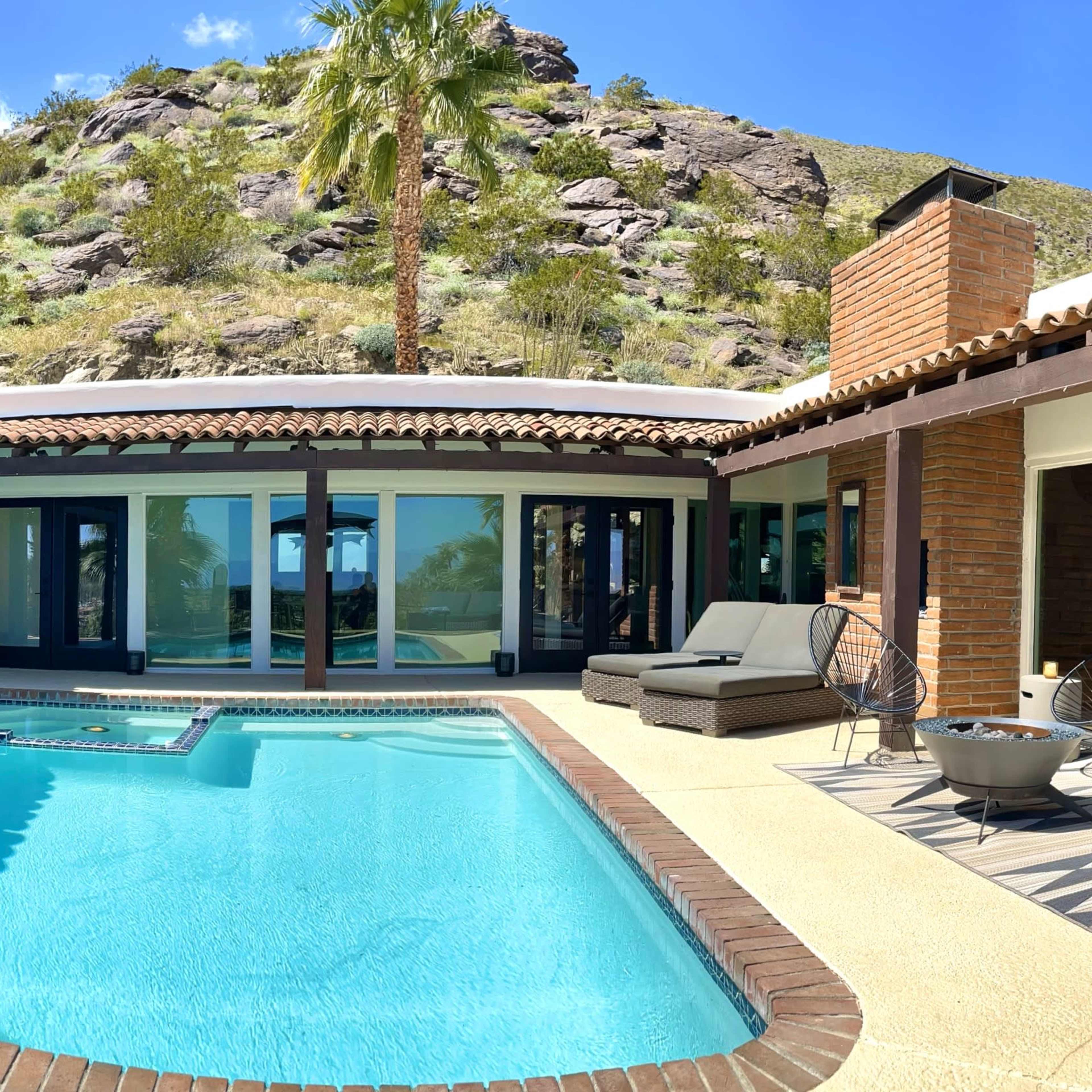 A modern home with a pool is surrounded by palm trees and a hillside, featuring large glass windows and a spacious patio area.