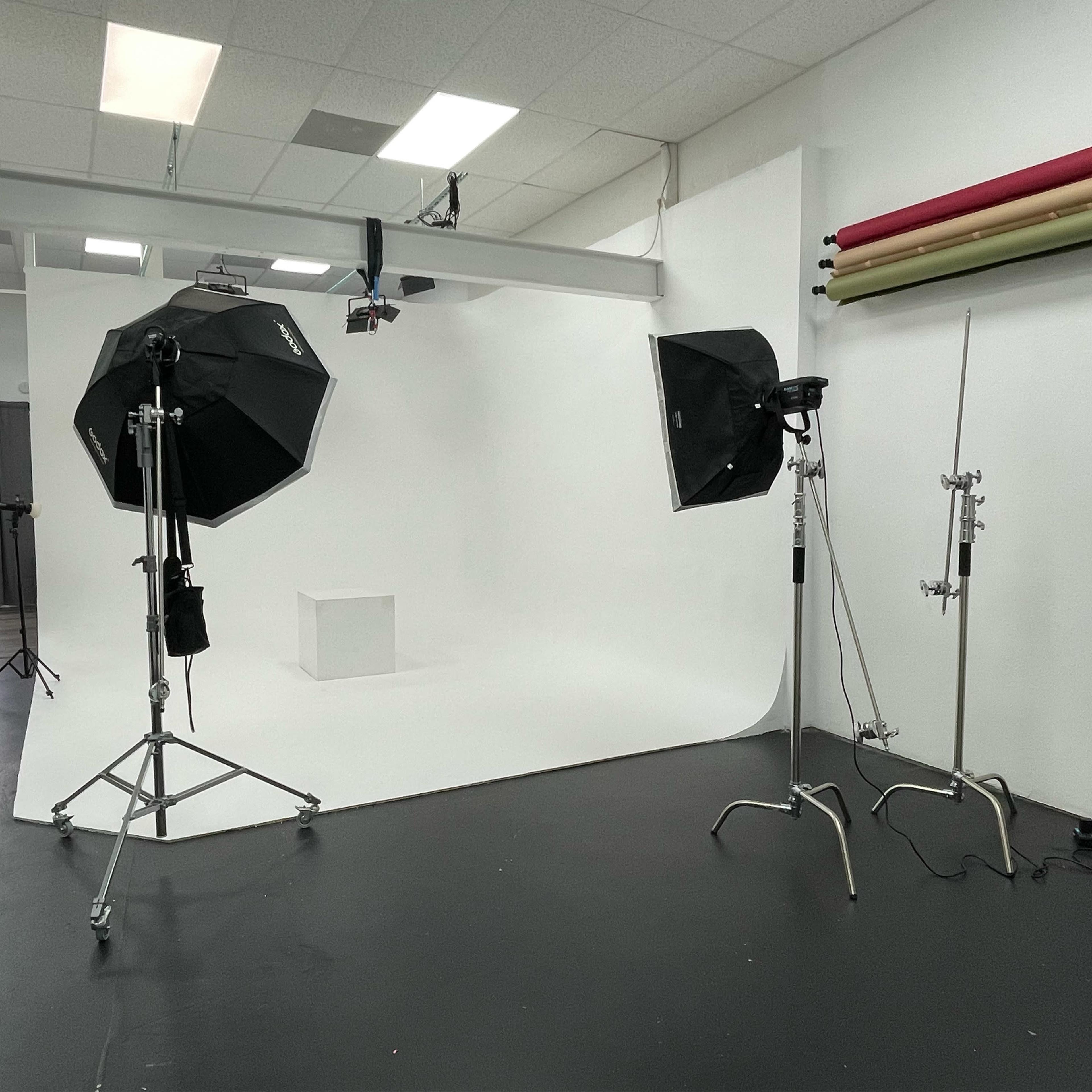 A photography studio with two light softboxes, a white backdrop, and various equipment, including a light stand and a reflector.