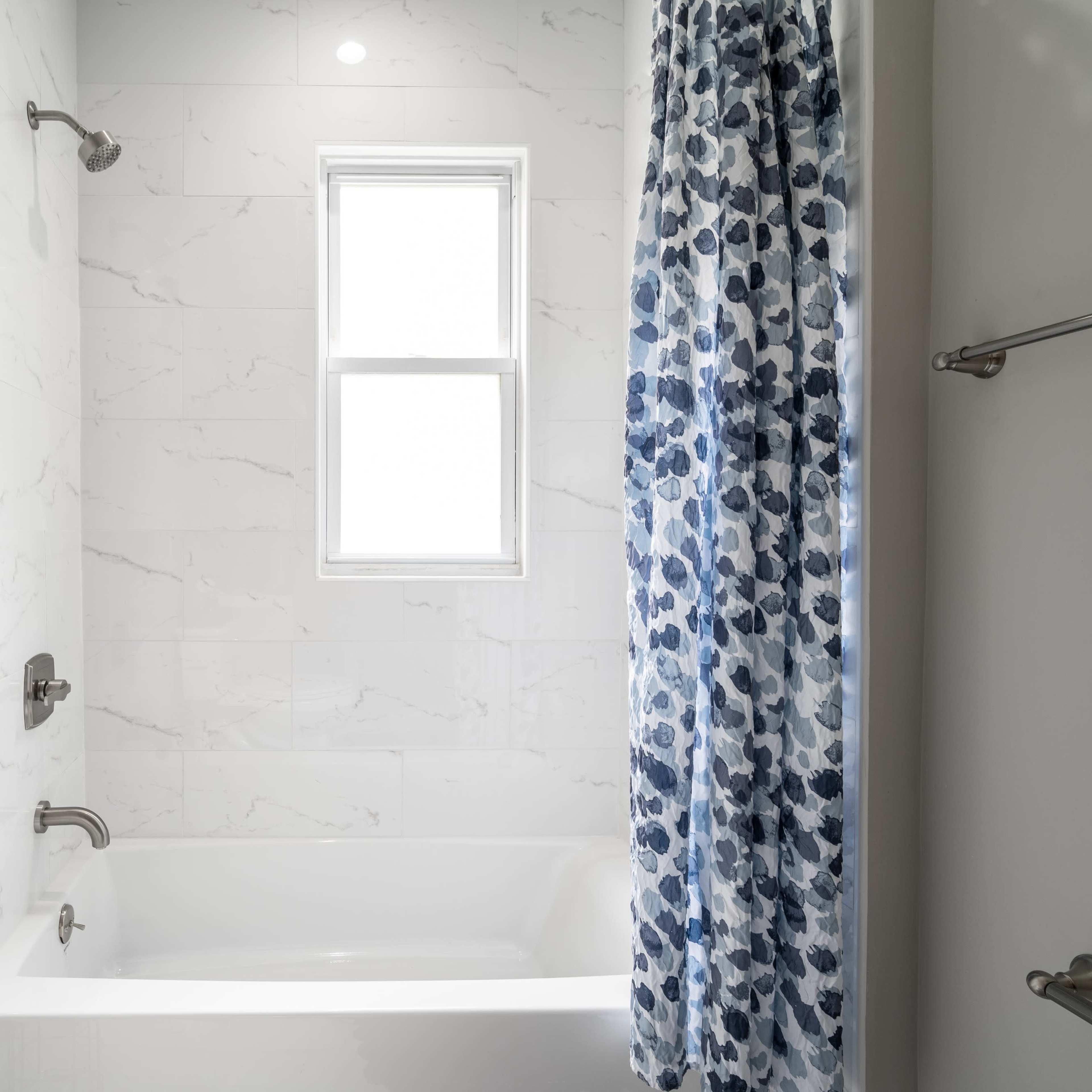 A modern bathroom with a white bathtub, marble-patterned walls, and a window covered by a blue floral shower curtain.