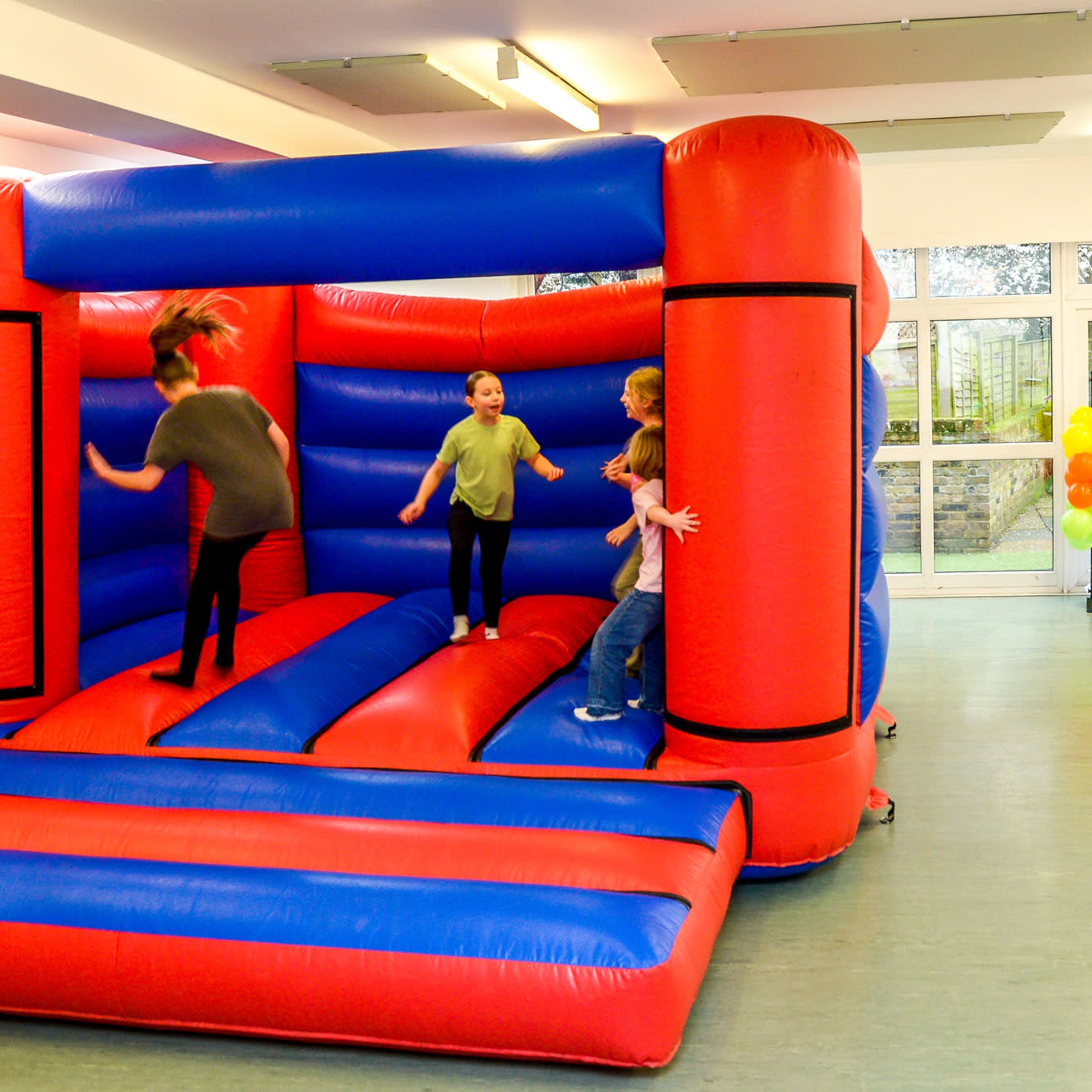 Children playing on a red and blue inflatable bouncer next to a decorated table with party supplies in a bright indoor space.