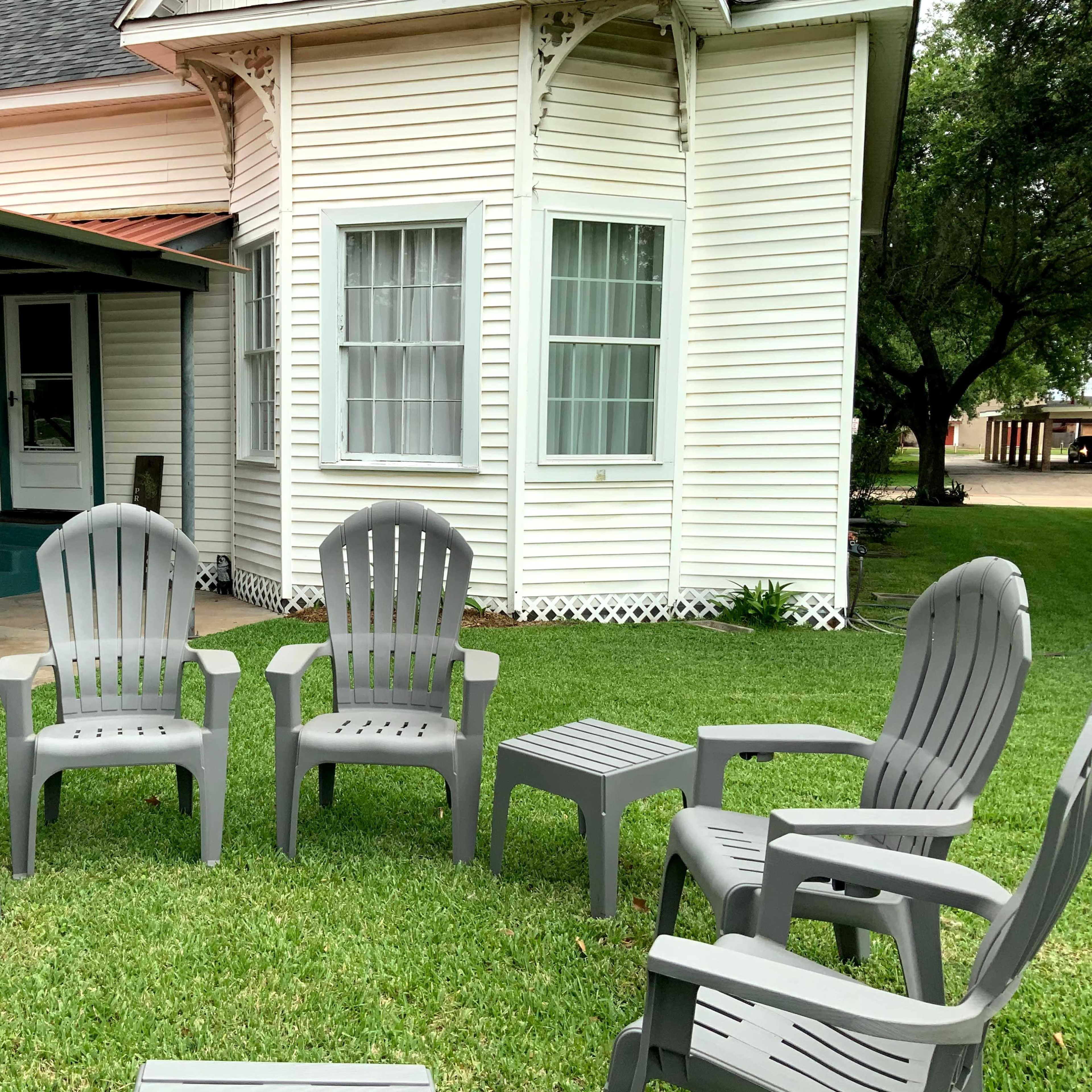 A set of gray plastic chairs is arranged in a circle on a grassy area in front of a white house.