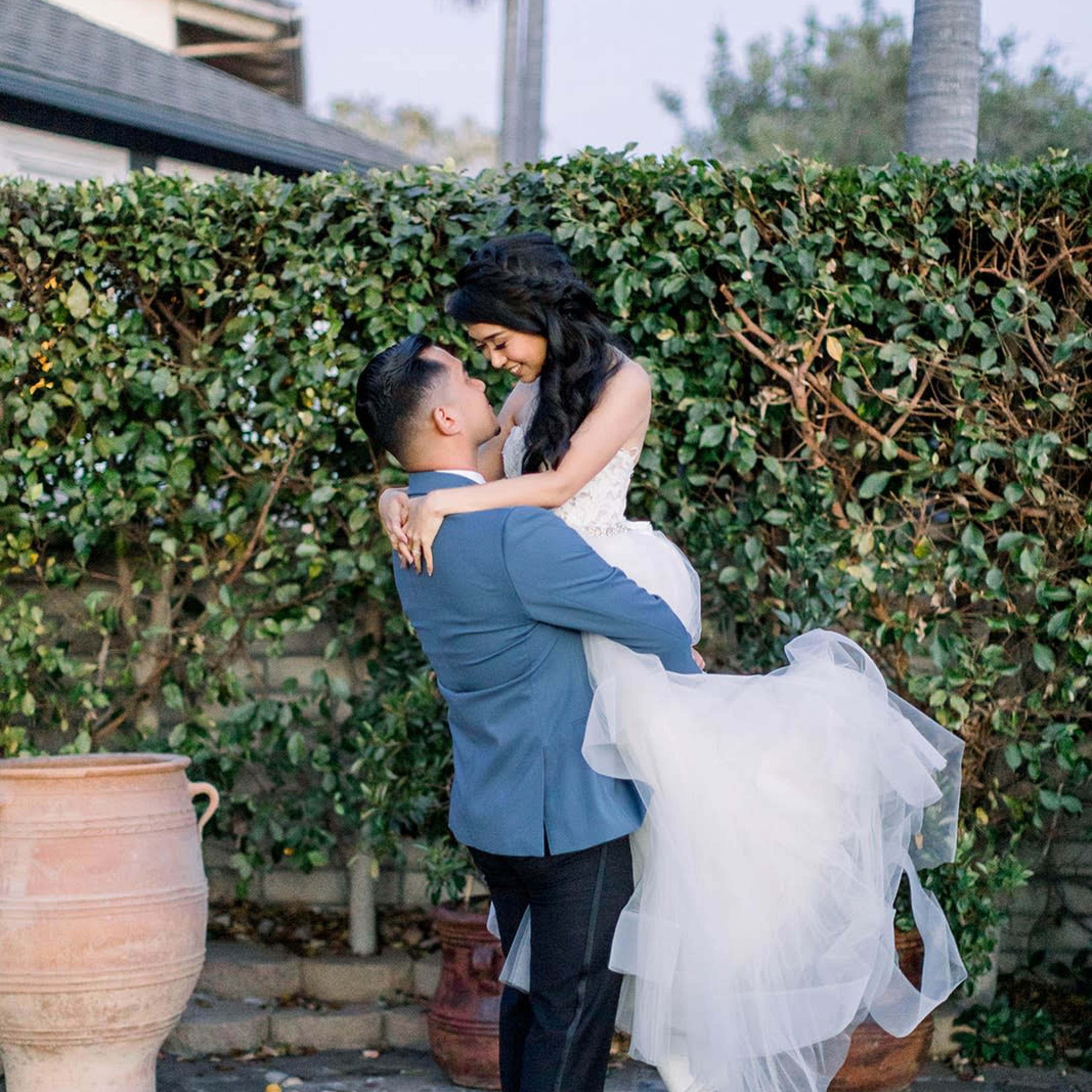 A groom lifts his bride in an outdoor setting with lush greenery and terra cotta pots.