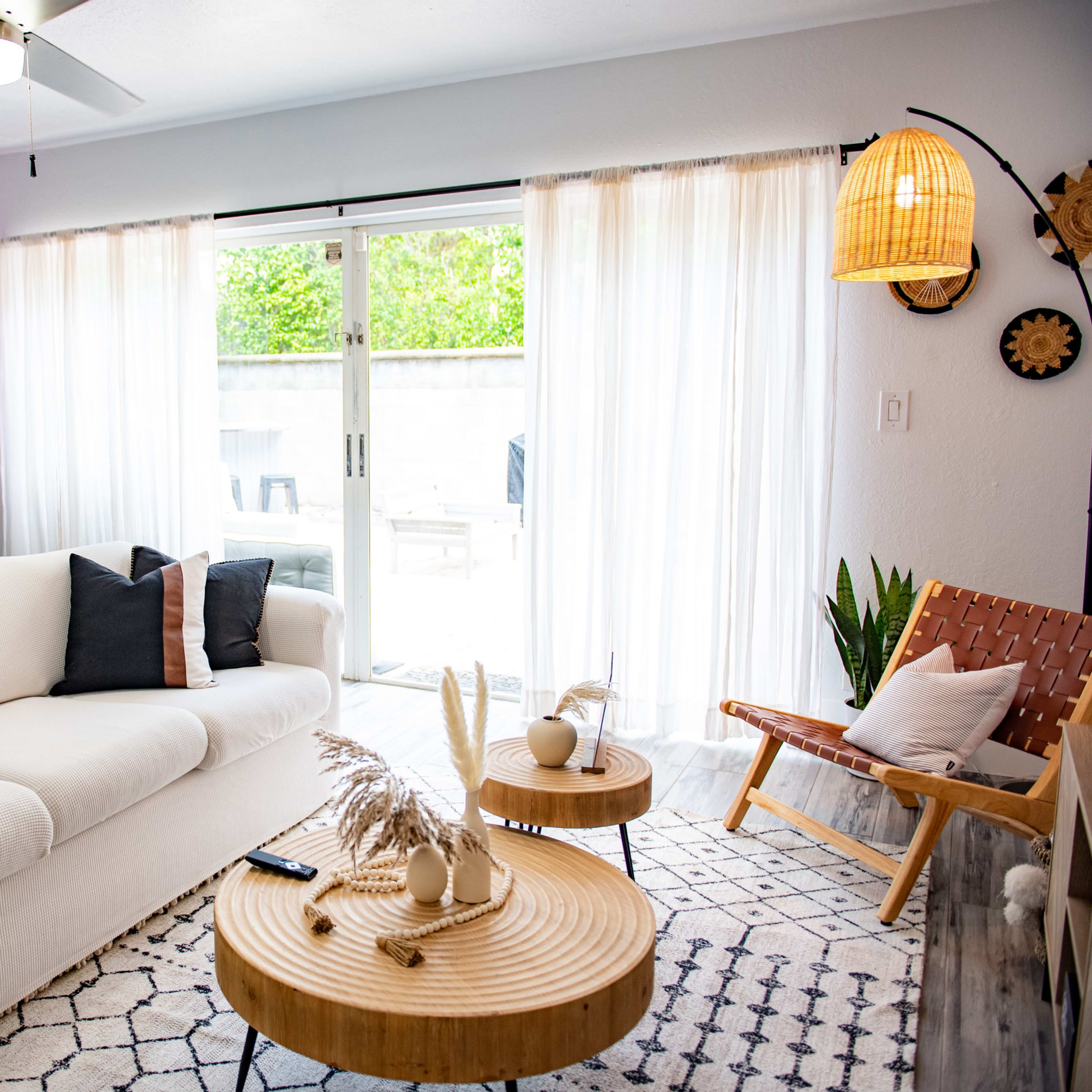 A bright living space featuring a white sofa with black and beige pillows, wooden coffee tables, and large glass doors leading to a patio, all decorated with plants and light fixtures.