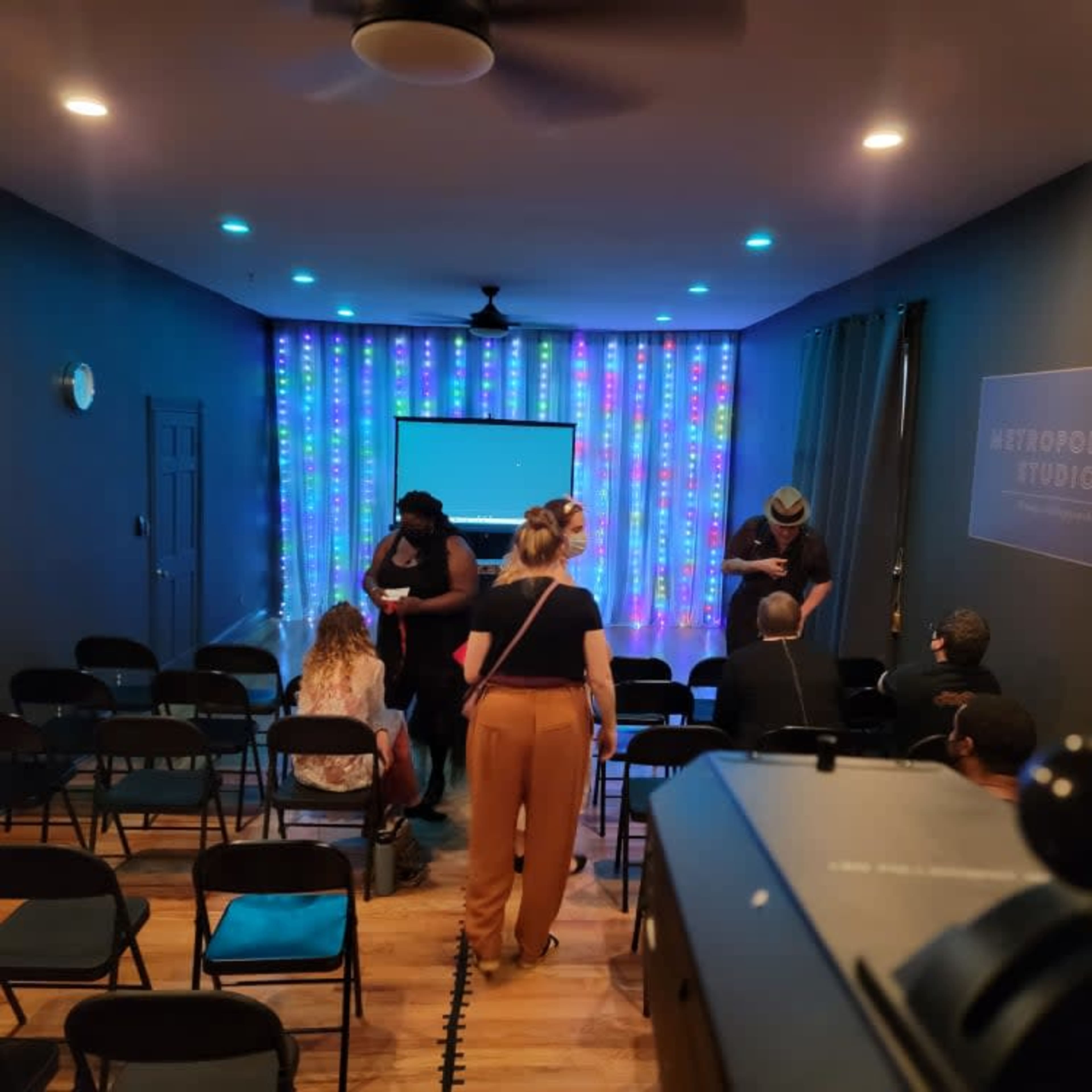 A group of people gathers in a small studio with colorful lights and a projector screen set up for a presentation or event.