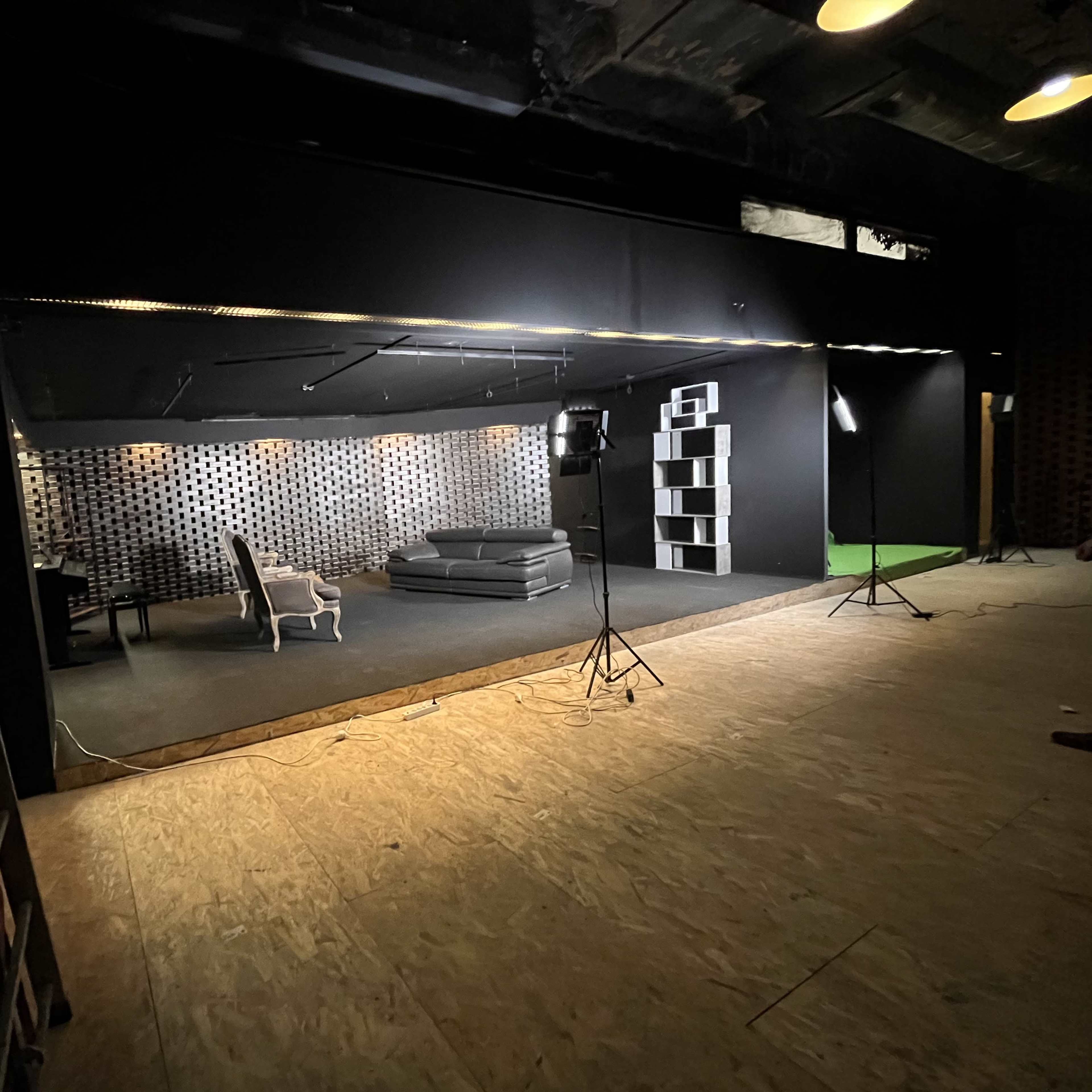A dimly lit studio with a black backdrop, featuring a gray sofa, a white chair, and a bookshelf against the wall, all set on a wooden floor.