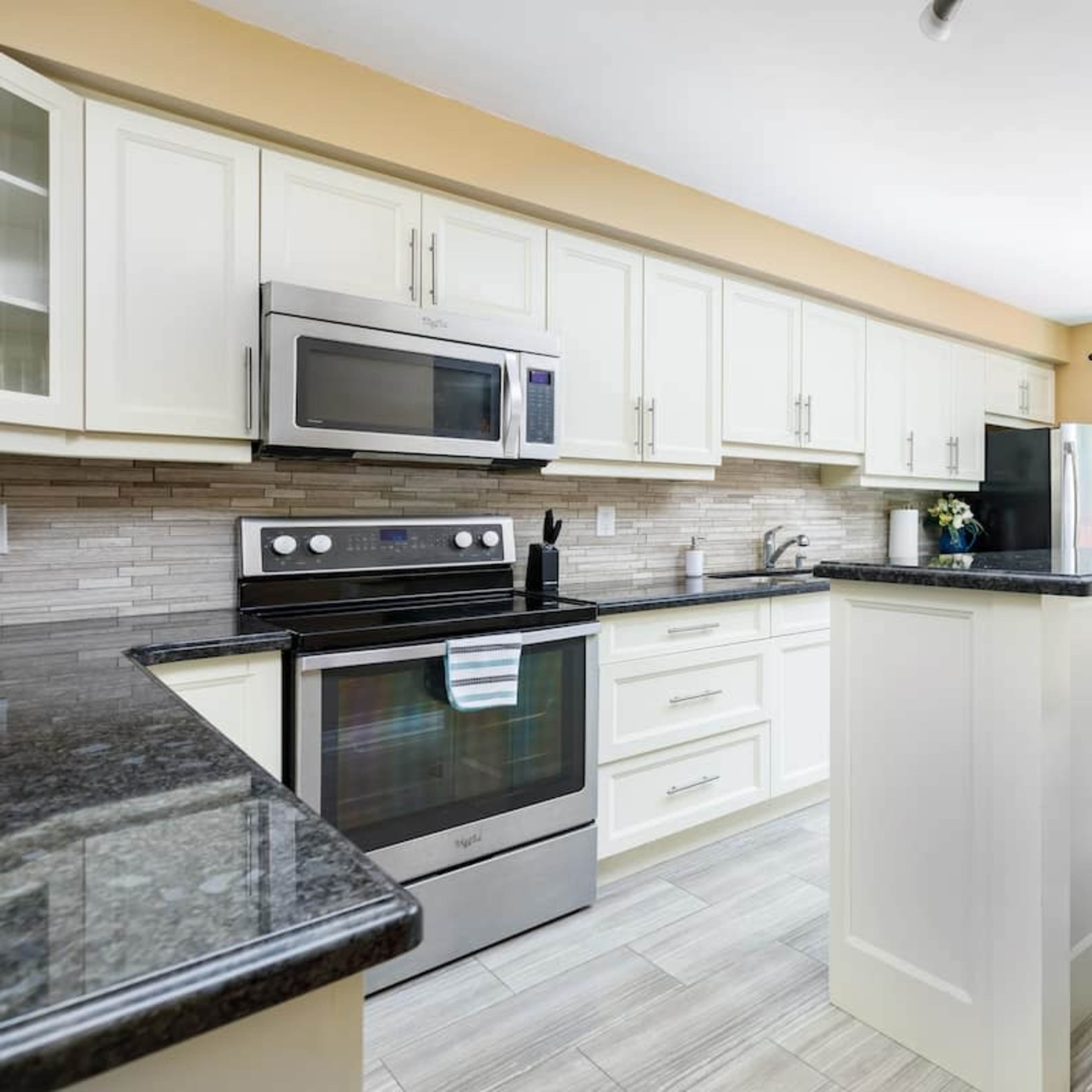 A modern kitchen features light cabinets, a dark countertop, stainless steel appliances, and a seating area with bar stools.